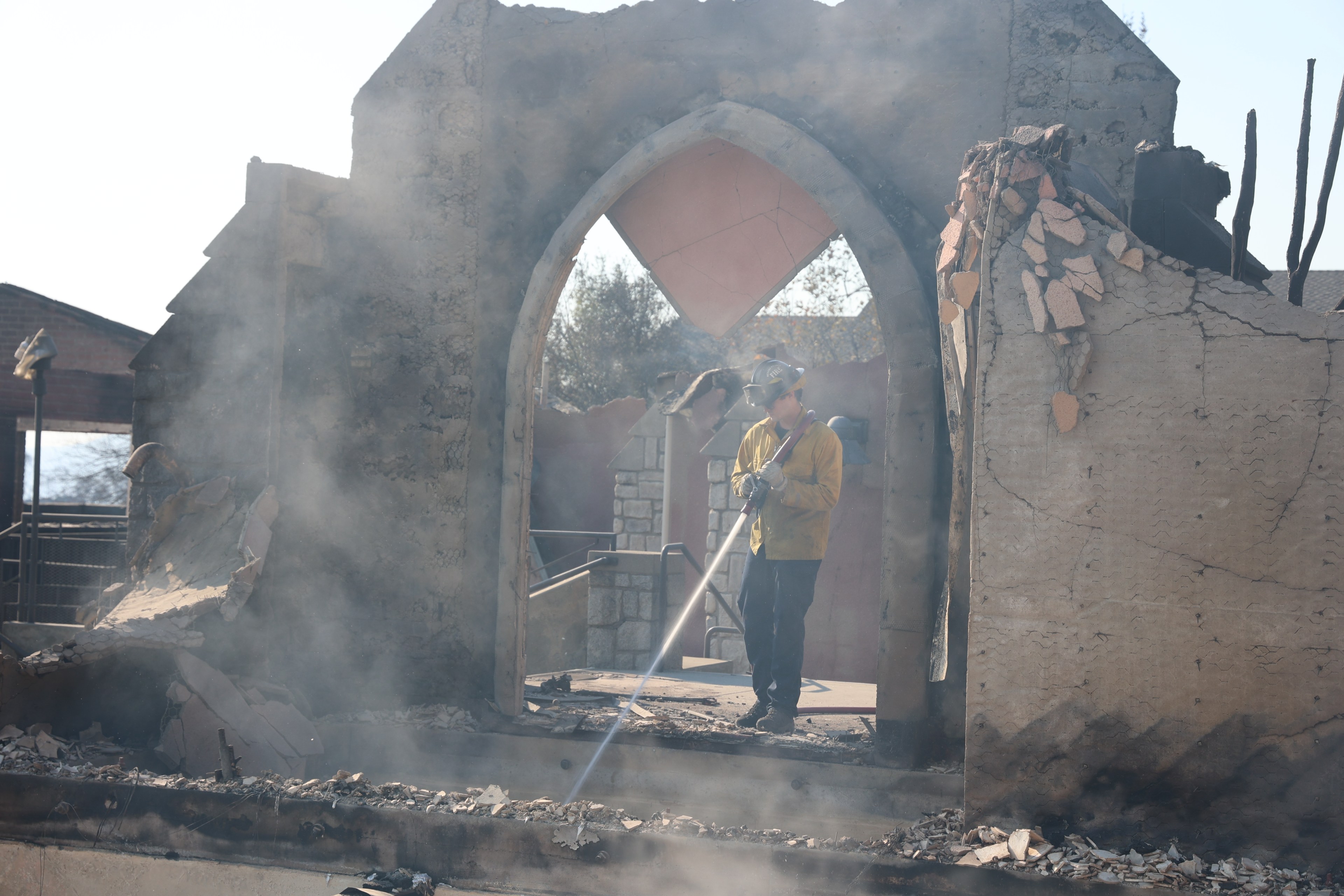 A firefighter in a yellow jacket is spraying water on a smoky, charred ruin through an arched window, surrounded by debris and crumbling walls.