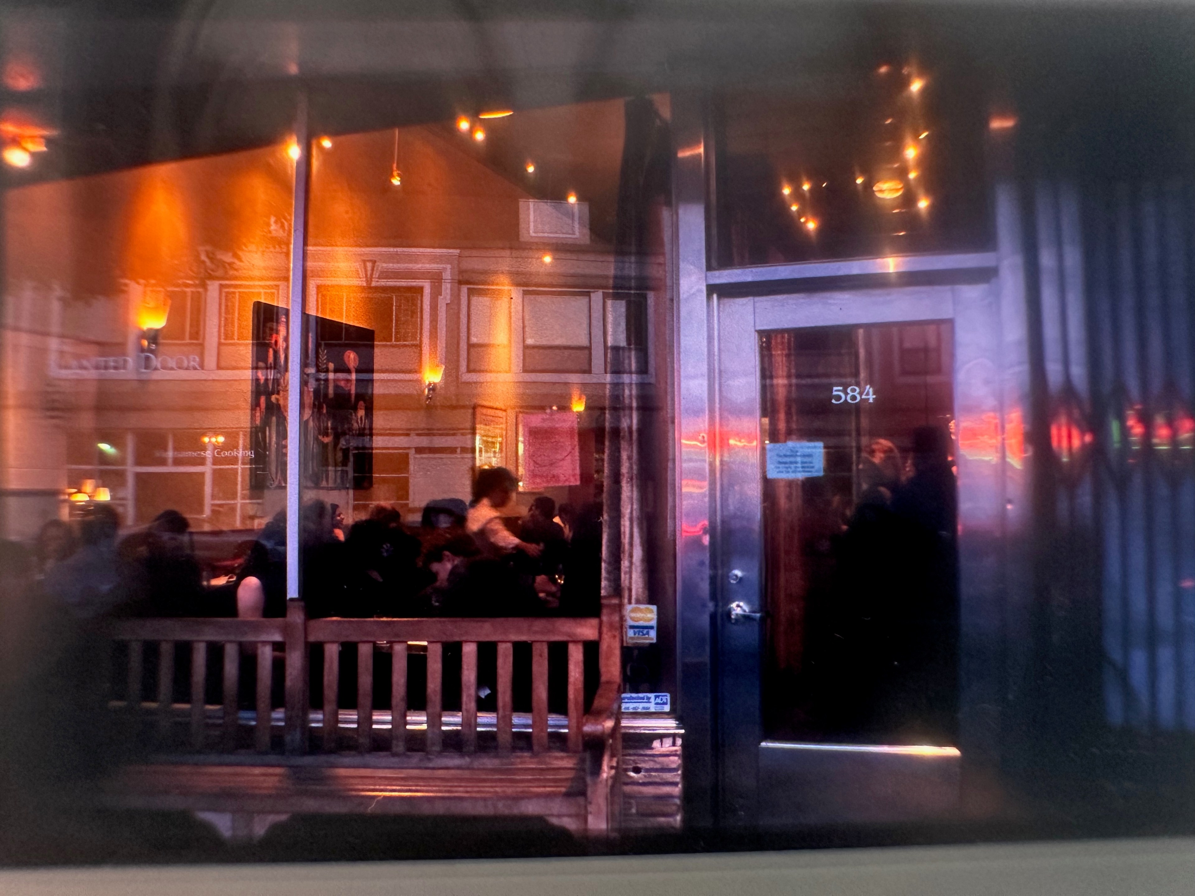 A busy, warmly lit restaurant interior with people seated inside. The glass reflects a building's facade and neon lights, with a door numbered 584.