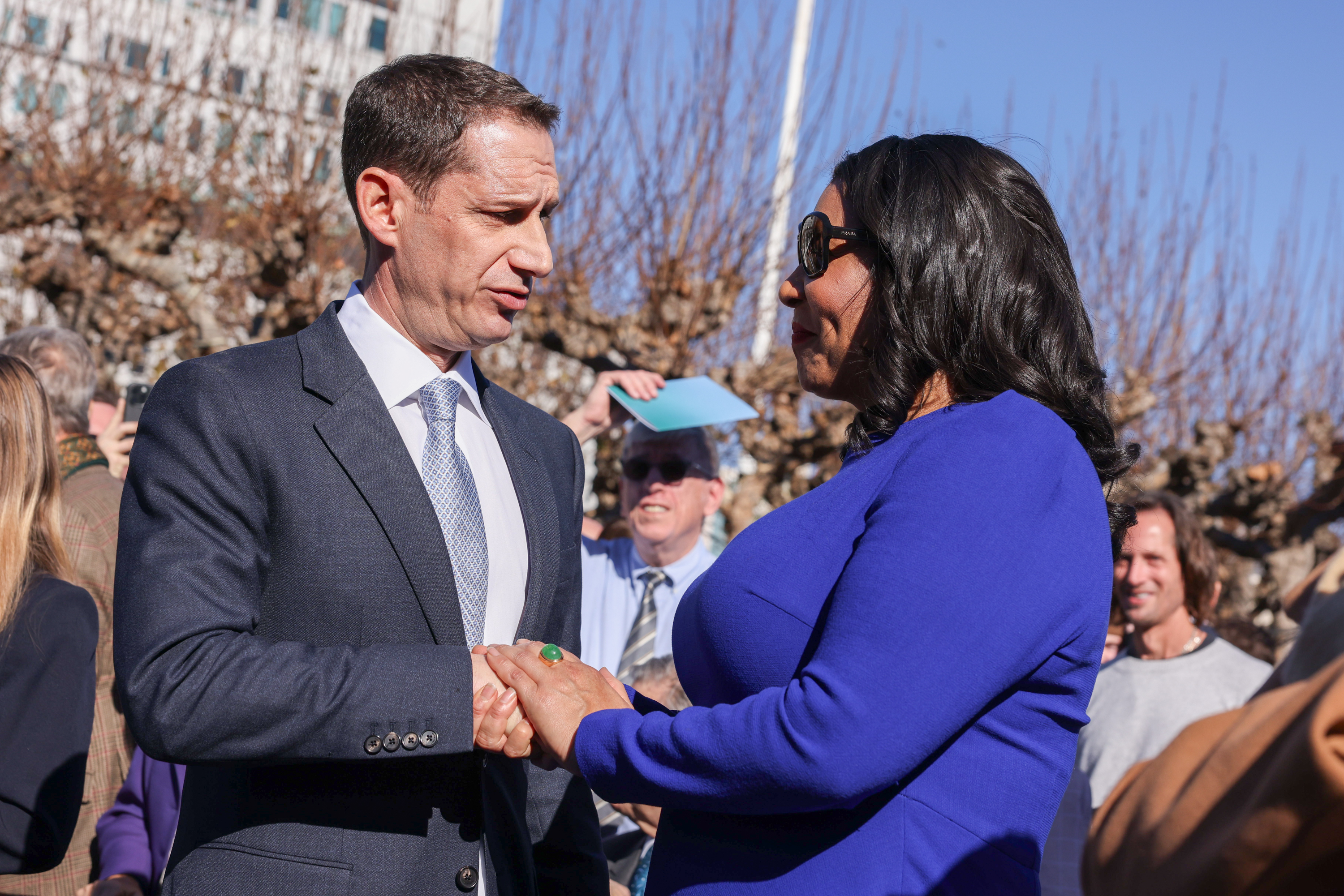 A man in a suit and a woman in a blue dress shake hands outdoors. They are surrounded by people, with a clear blue sky in the background.