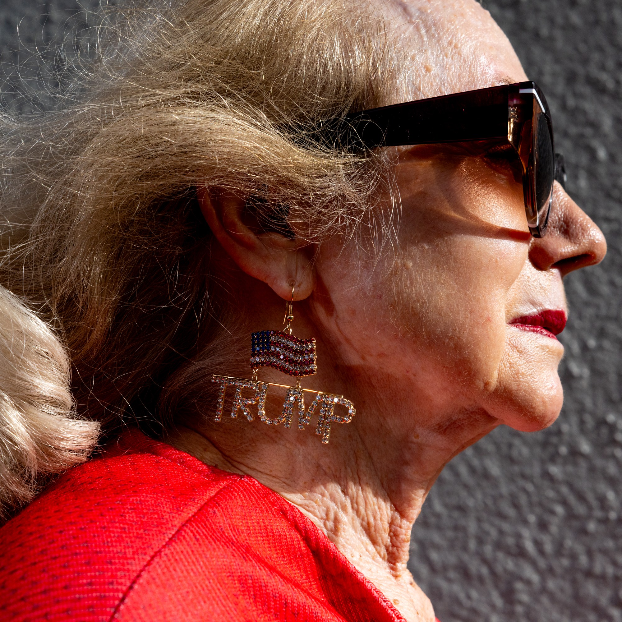 An elderly woman with sunglasses and blonde hair is wearing a red top and an earring featuring an American flag and the word "TRUMP."