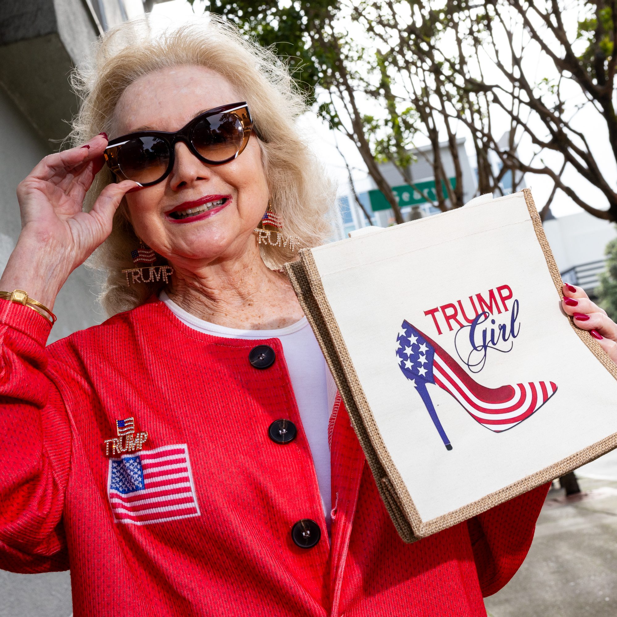 An older woman in a red outfit, wearing sunglasses, smiles while holding a bag that reads &quot;Trump Girl&quot; with a high heel in American flag colors.