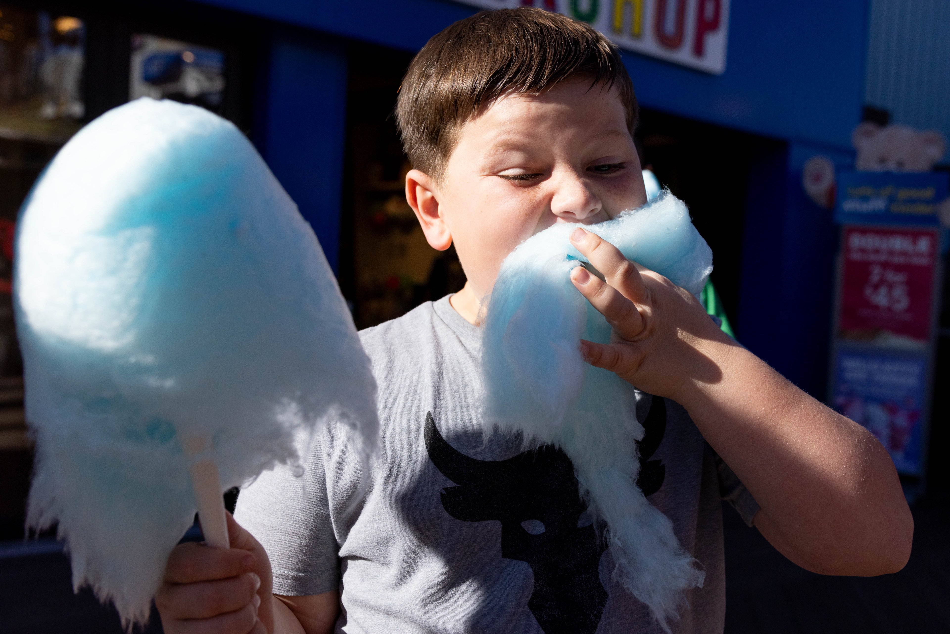 A child in a gray shirt eagerly eats blue cotton candy, holding two large sticks of it. They are standing in front of a colorful shop sign.