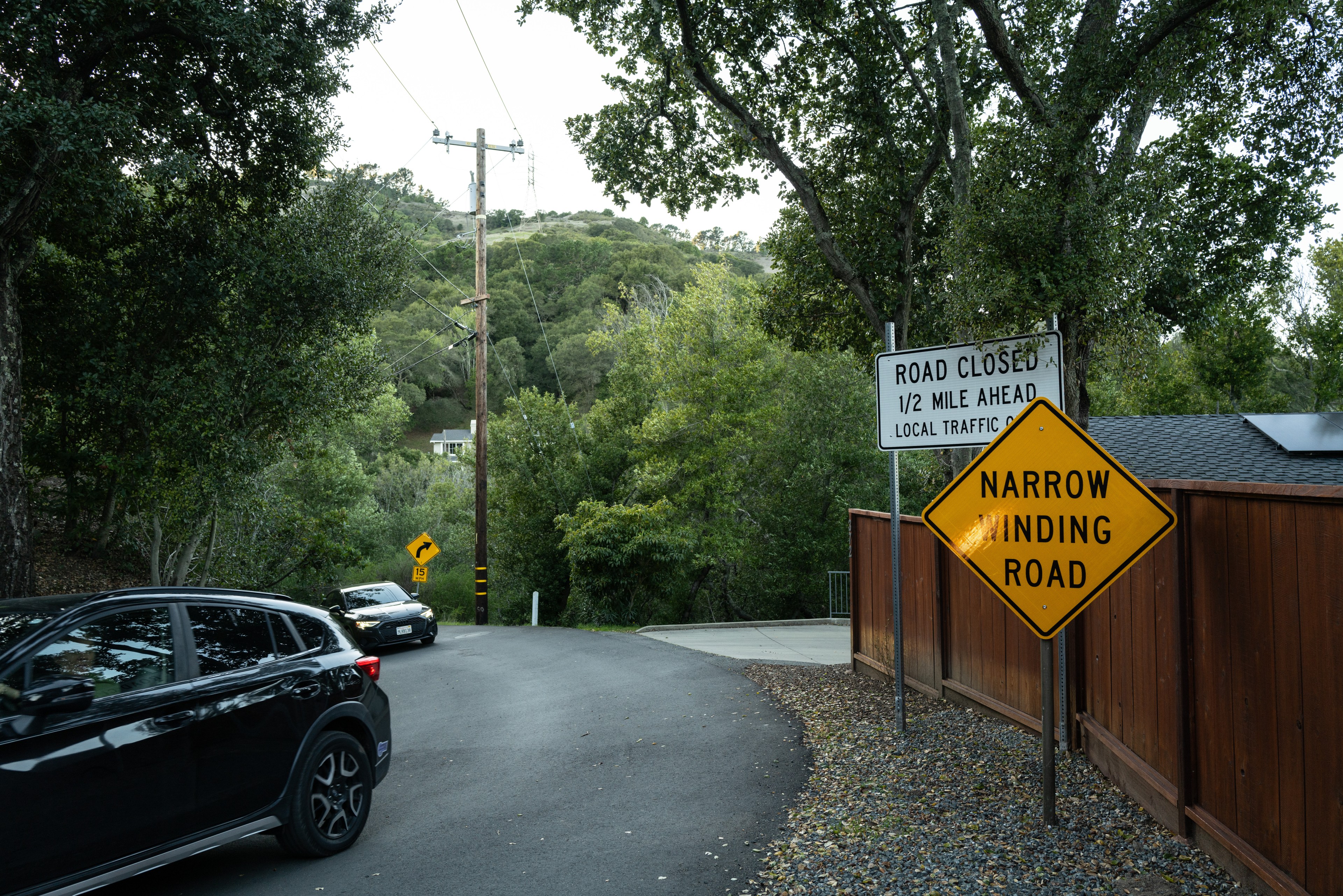 A winding road through trees with a yellow &quot;Narrow Winding Road&quot; sign and a white &quot;Road Closed 1/2 Mile Ahead&quot; sign. Two cars are on the road.