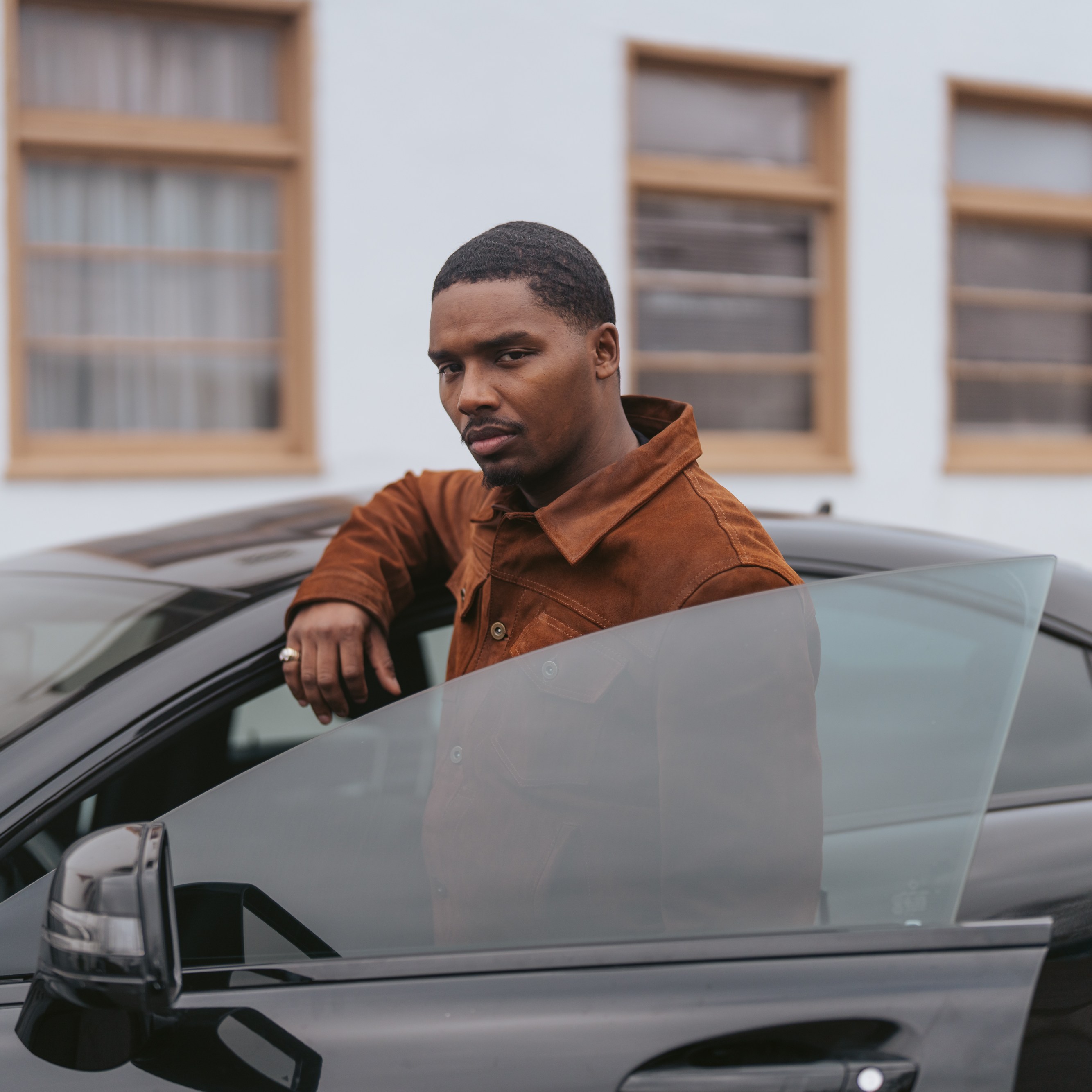 A person in a brown jacket leans on the open door of a black car, facing the camera. The background shows a building with several windows.