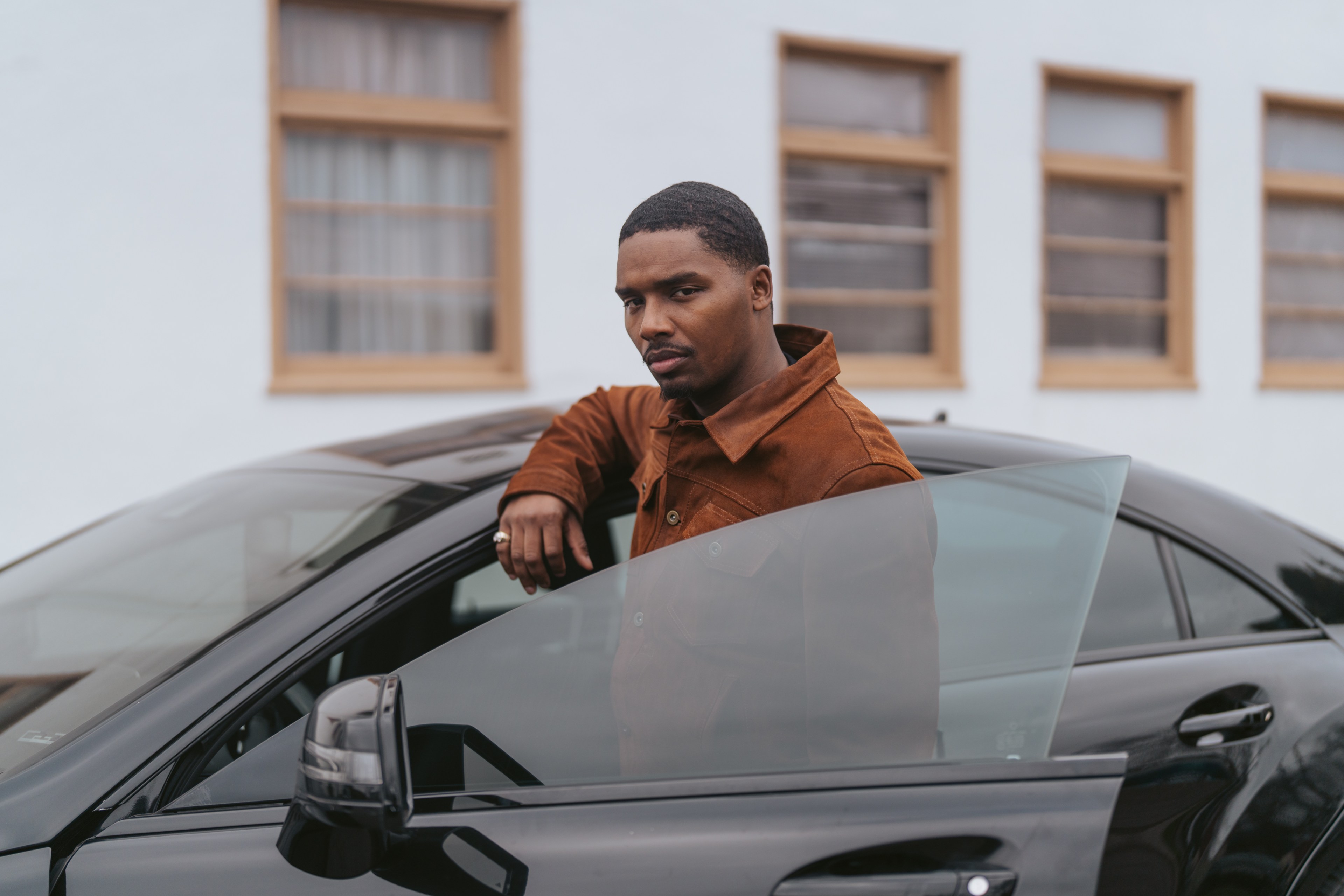 A person in a brown jacket leans on the open door of a black car, facing the camera. The background shows a building with several windows.