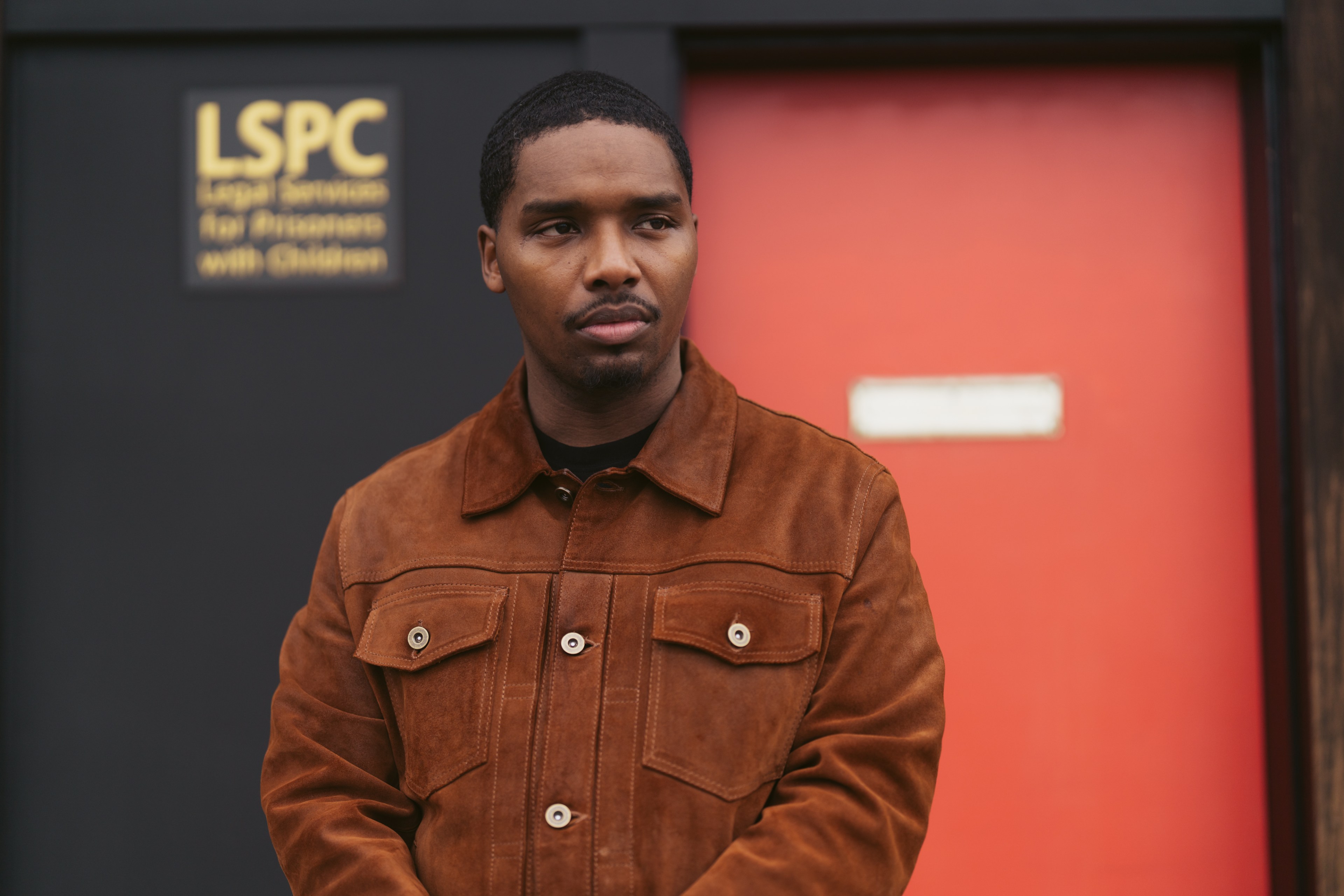 A man in a brown jacket stands in front of a black wall with a sign that says &quot;LSPC Legal Services for Prisoners with Children,&quot; and a red door is behind him.