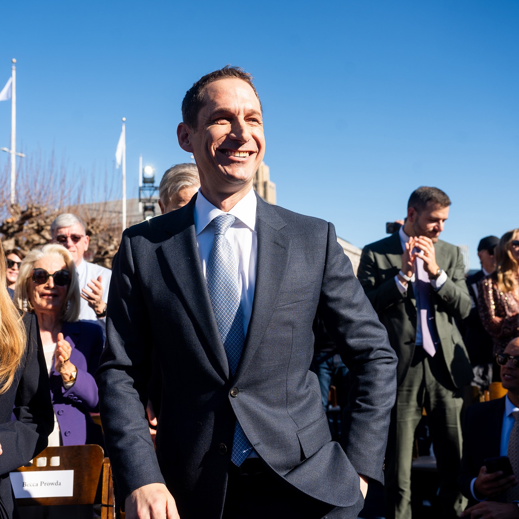 A man in a suit smiles broadly among a crowd outdoors. People are clapping, and there's a clear blue sky overhead. A woman with long hair is next to him.