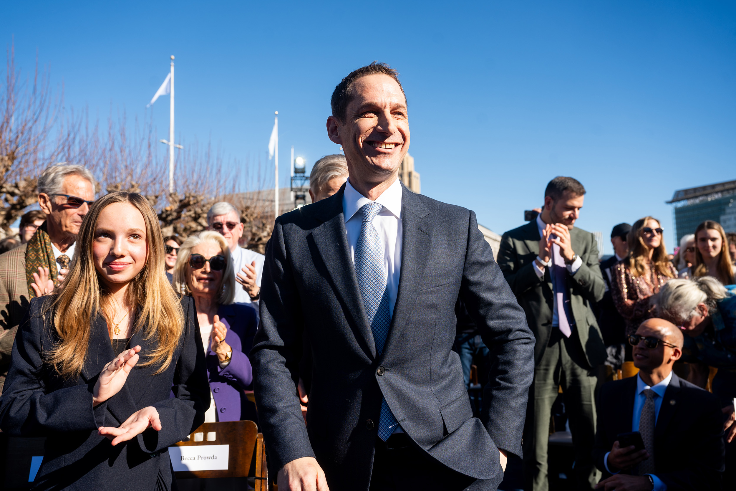 A man in a suit smiles broadly among a crowd outdoors. People are clapping, and there's a clear blue sky overhead. A woman with long hair is next to him.