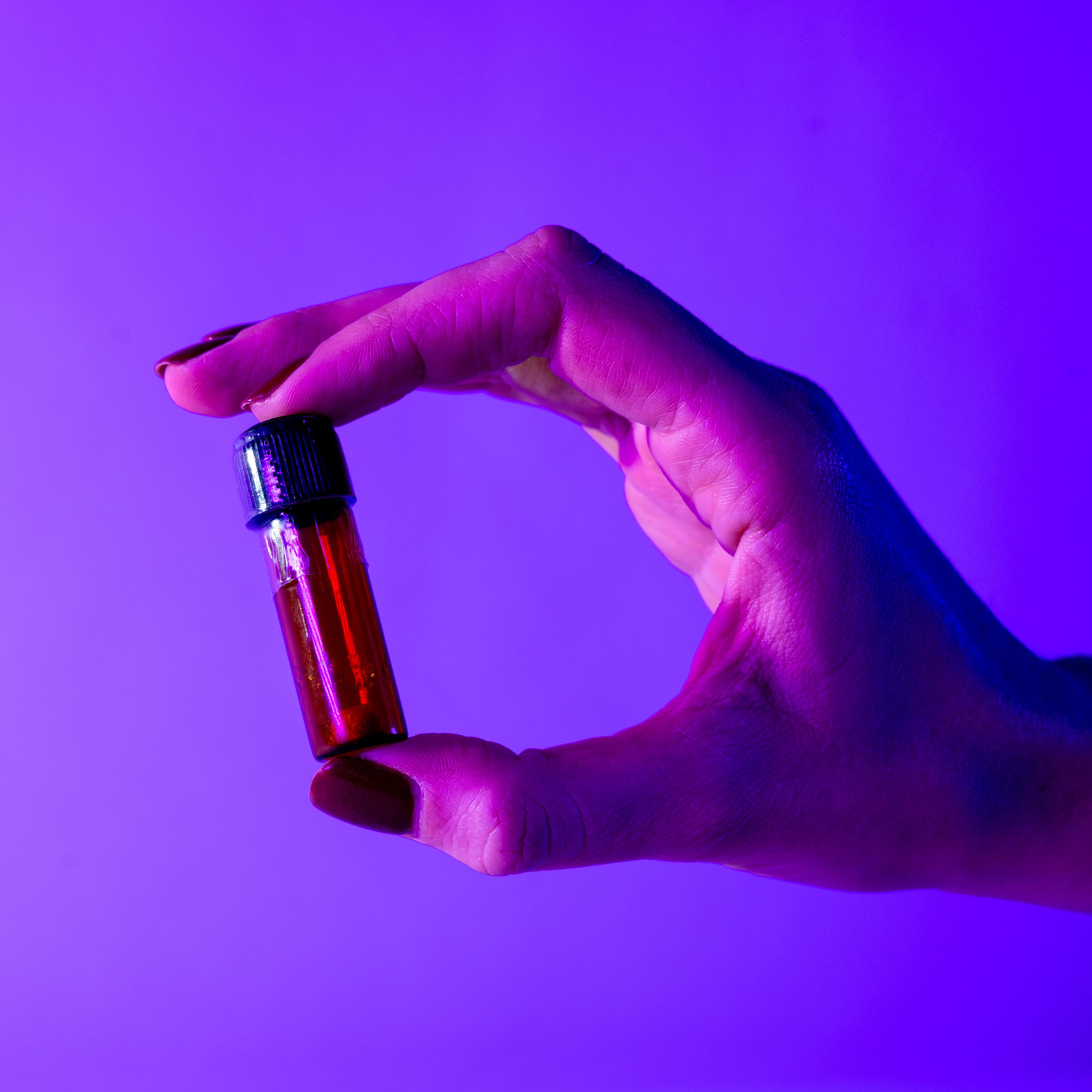 A hand is holding a small glass vial with red liquid, under purple lighting. The vial has a black cap, and the background is a gradient of blue and purple.