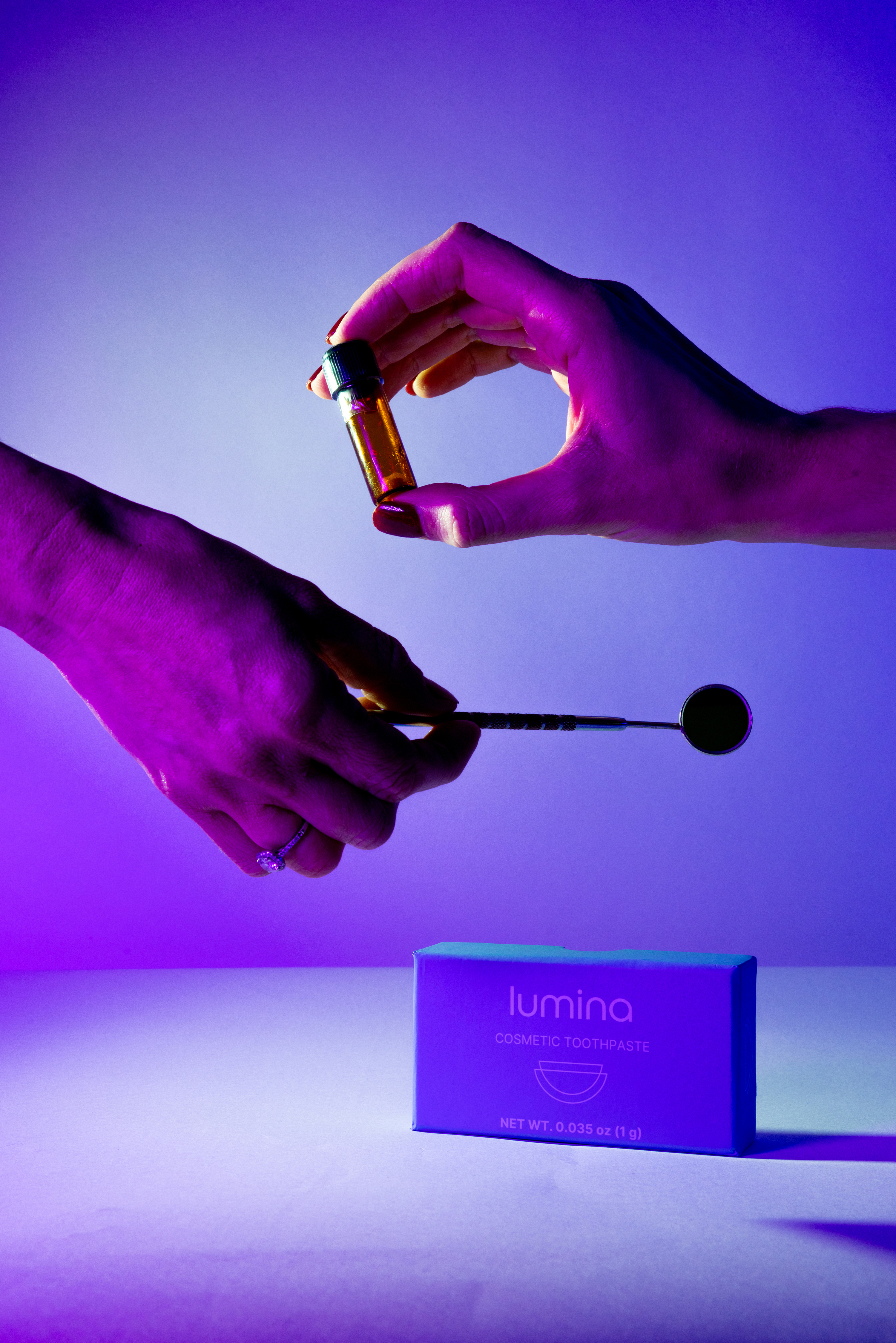 Two hands exchange a small brown vial against a purple-lit background. Below, a blue box labeled &quot;Lumina Cosmetic Toothpaste&quot; rests on a surface.