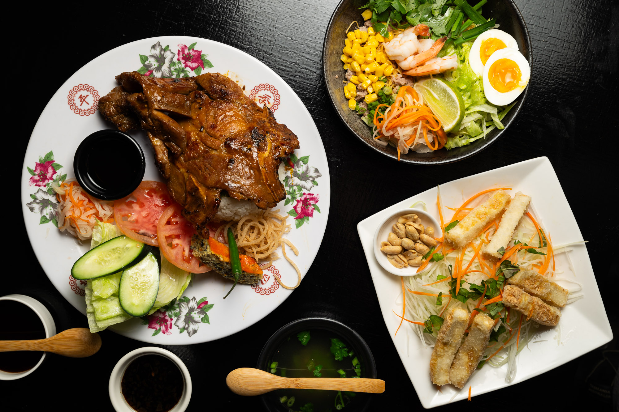 The image shows a table with a grilled meat dish, a noodle salad with fried fish, and a bowl with shrimp, corn, and eggs, accompanied by sauces and soup.