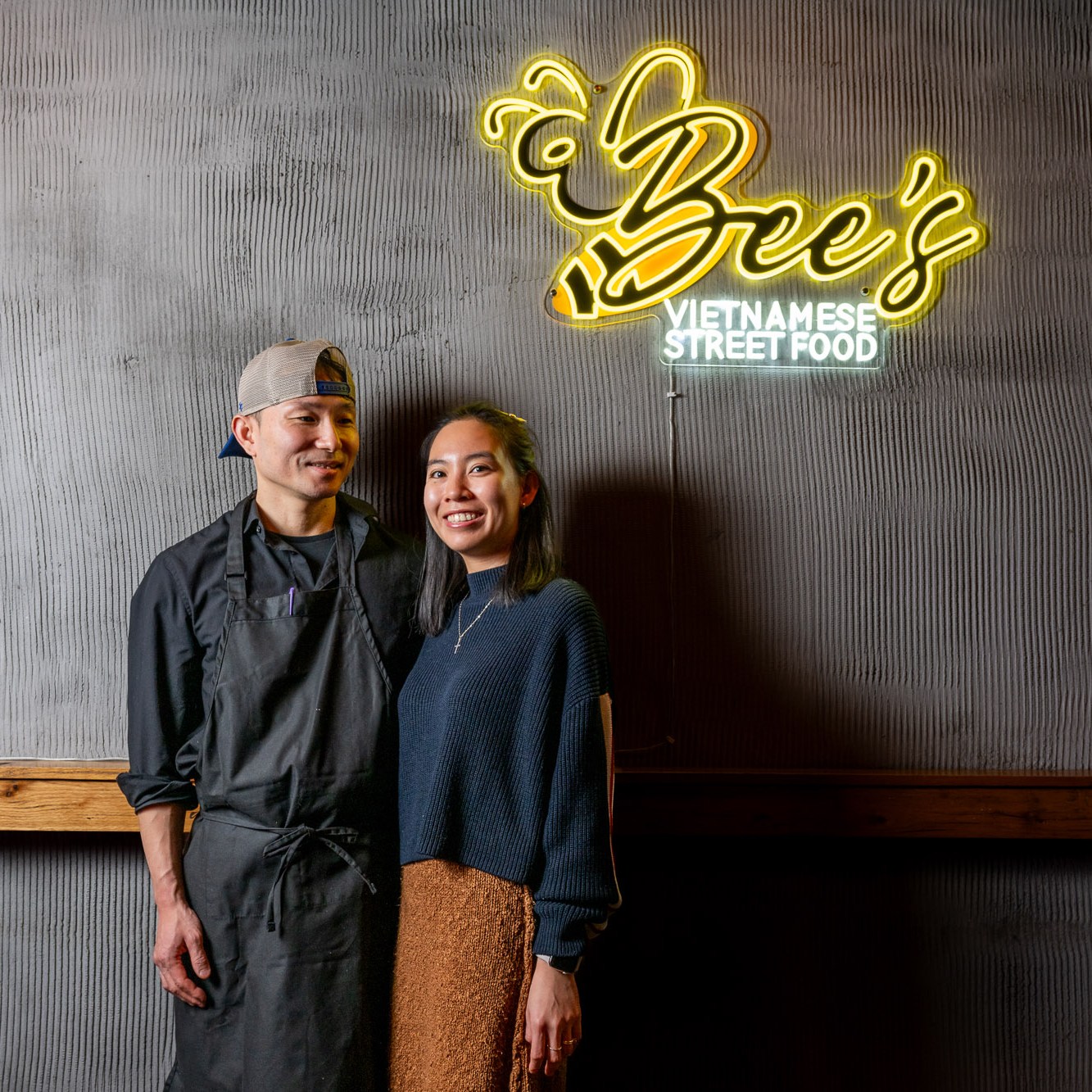 A man in a chef's uniform and a woman stand smiling under a neon sign that reads "Bee's Vietnamese Street Food" on a textured wall.