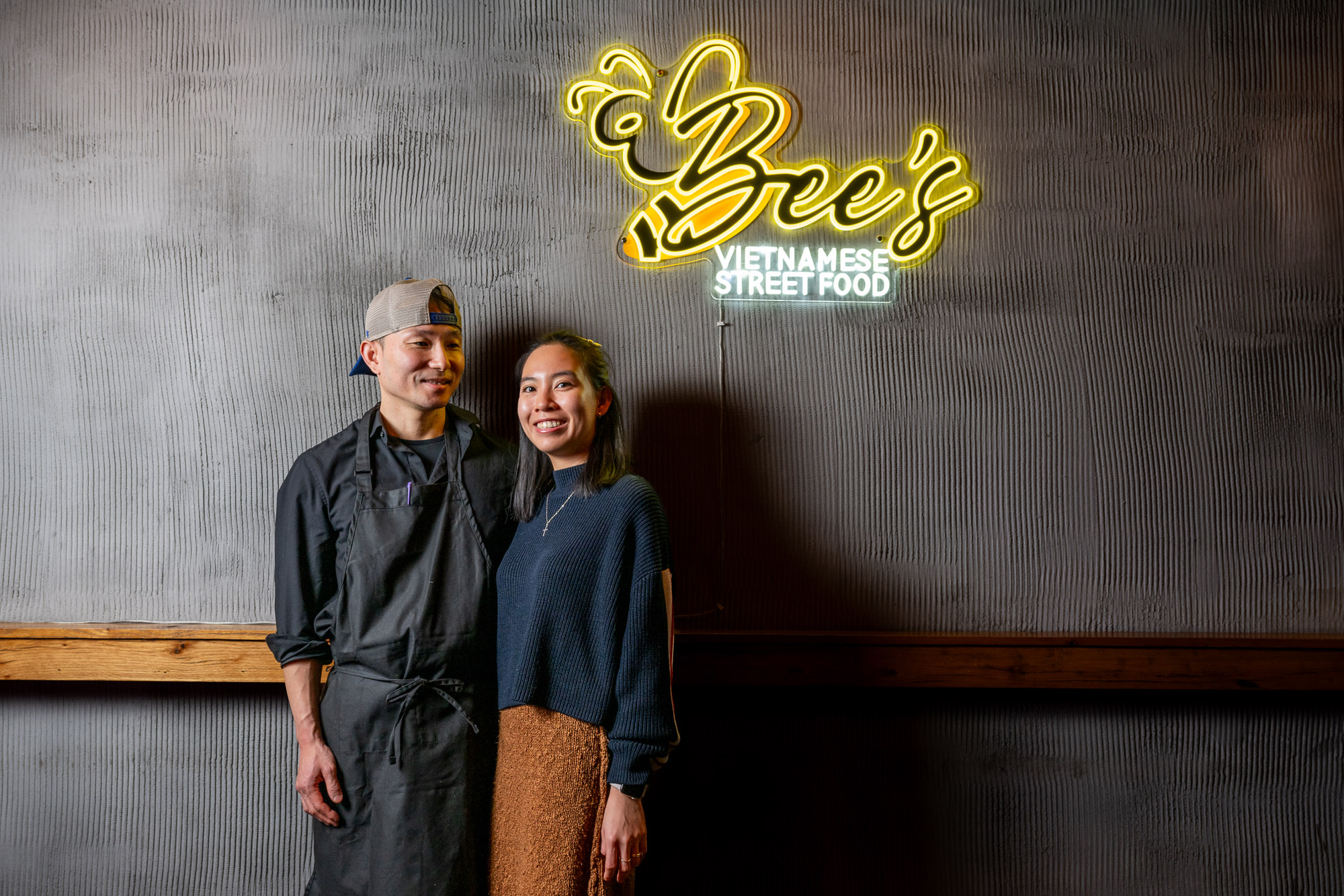 A man in a chef's uniform and a woman stand smiling under a neon sign that reads "Bee's Vietnamese Street Food" on a textured wall.