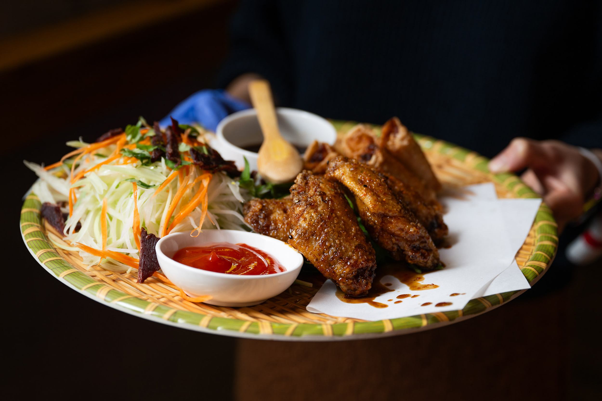 A plate holds crispy chicken wings, fresh shredded vegetables, spring rolls, and small bowls of red sauce and dark sauce, all on a woven tray.