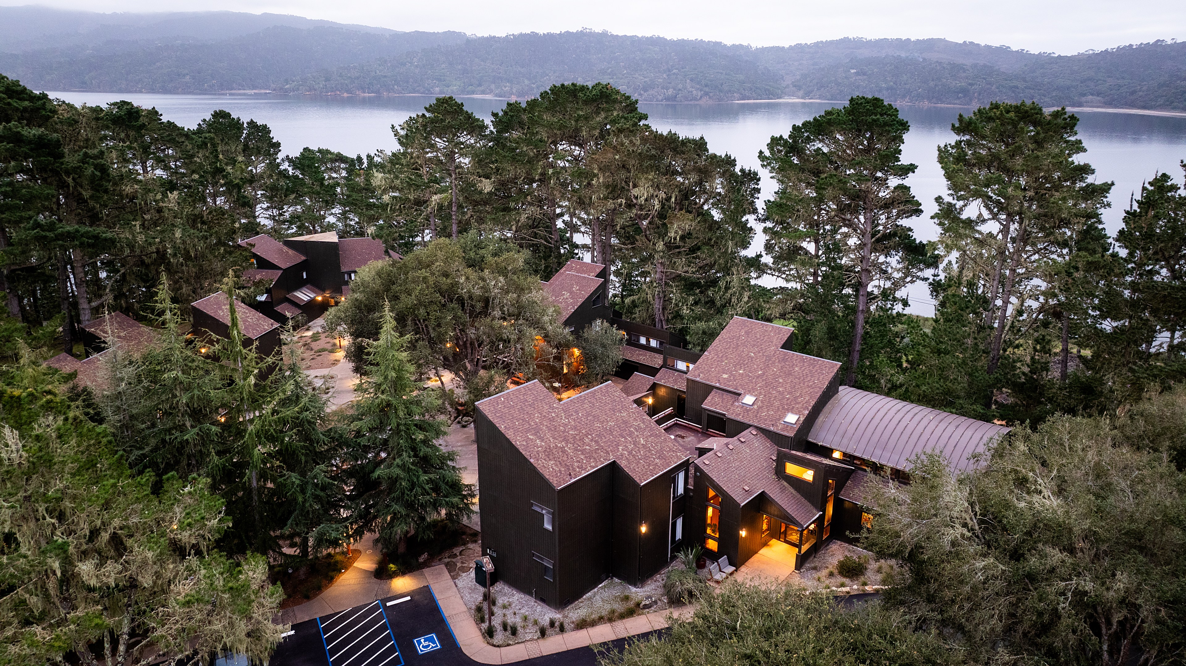 A cluster of modern buildings with brown roofs is nestled among tall trees by a serene lake, with a backdrop of forested hills and a slightly cloudy sky.