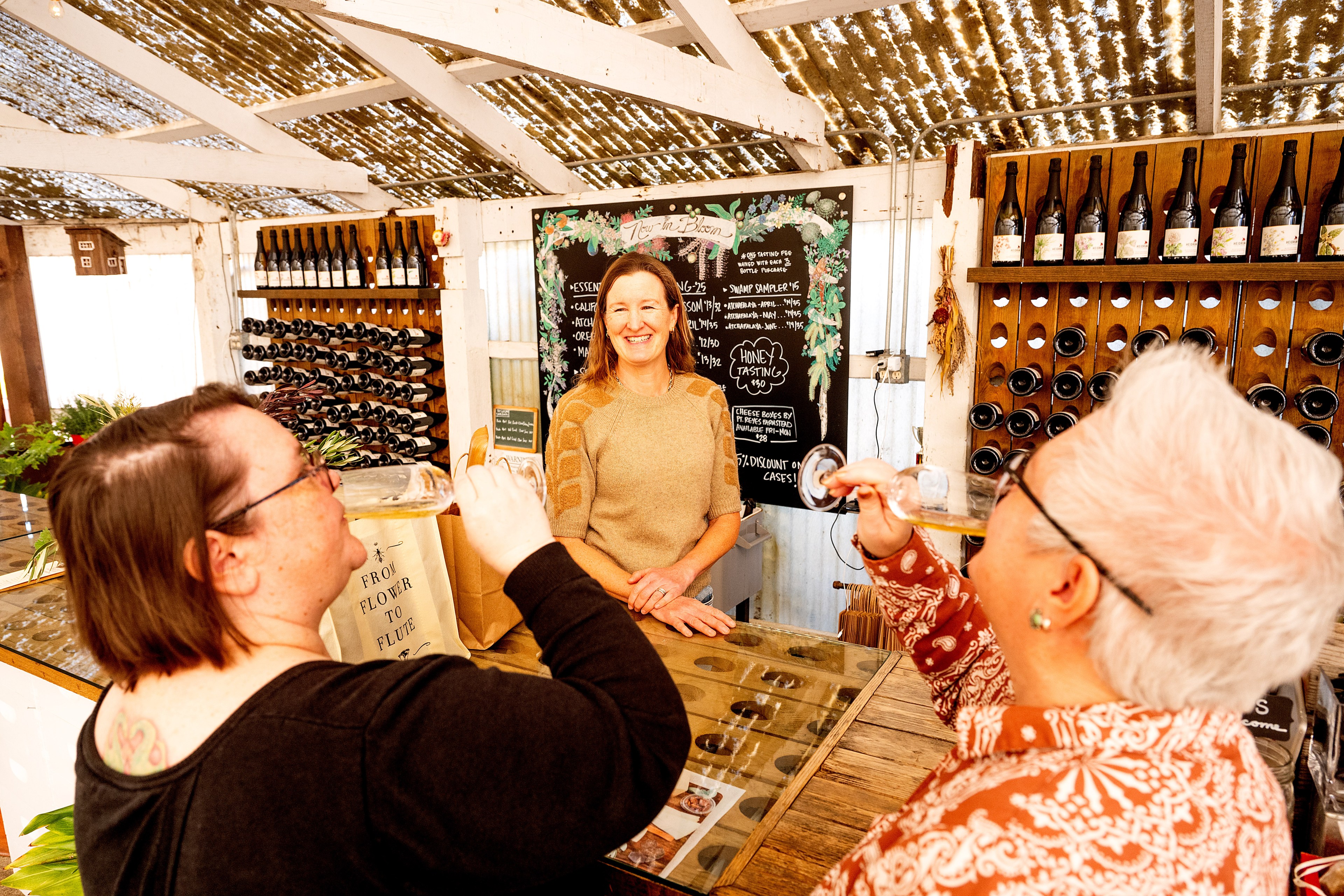 A smiling woman stands at a counter with wine bottles behind her, while two women sip from glasses in front of her. There's a chalkboard displaying options.