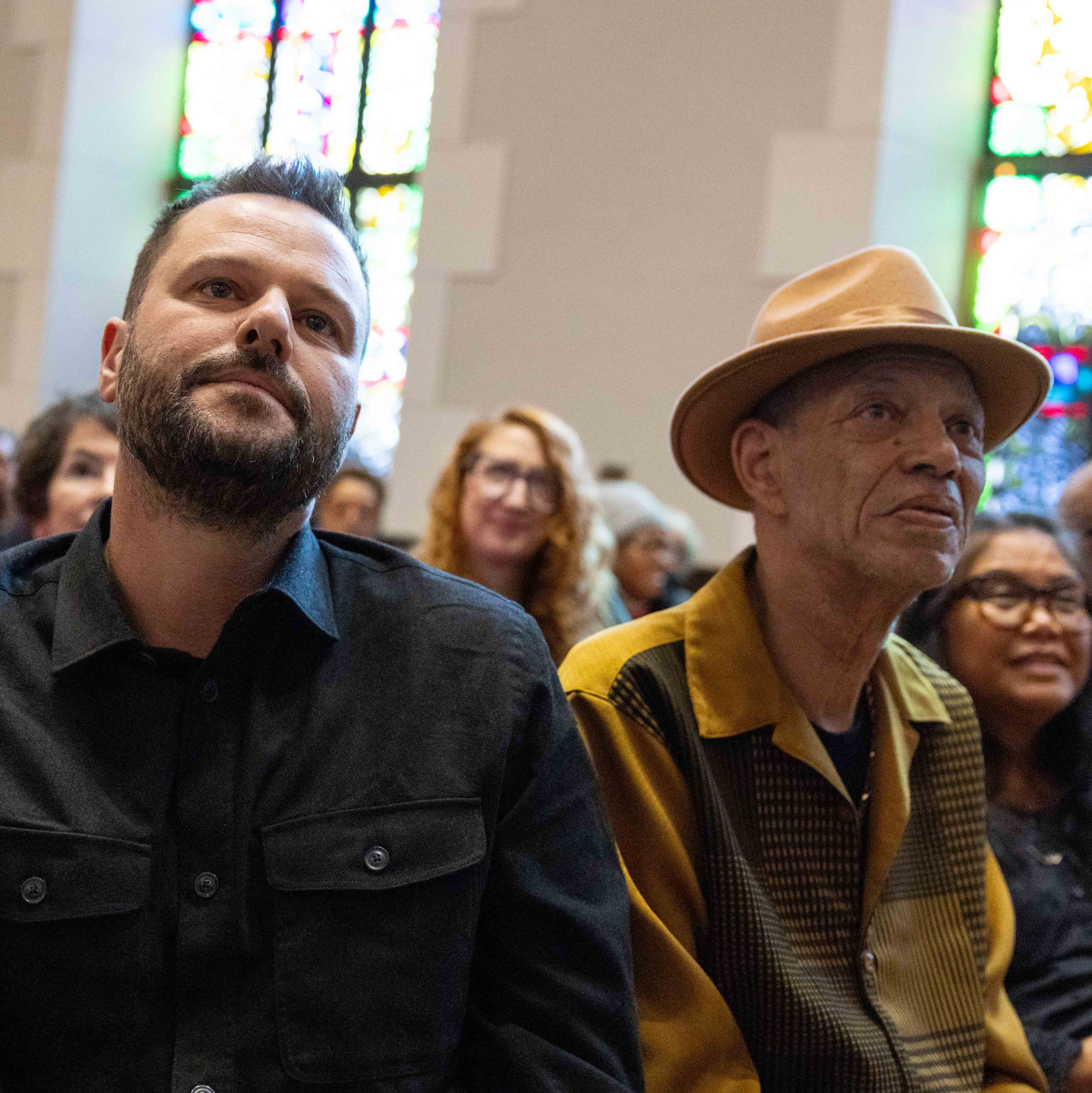 People are seated in a church, facing forward with expressions of interest and thoughtfulness. Stained glass windows are visible in the background.