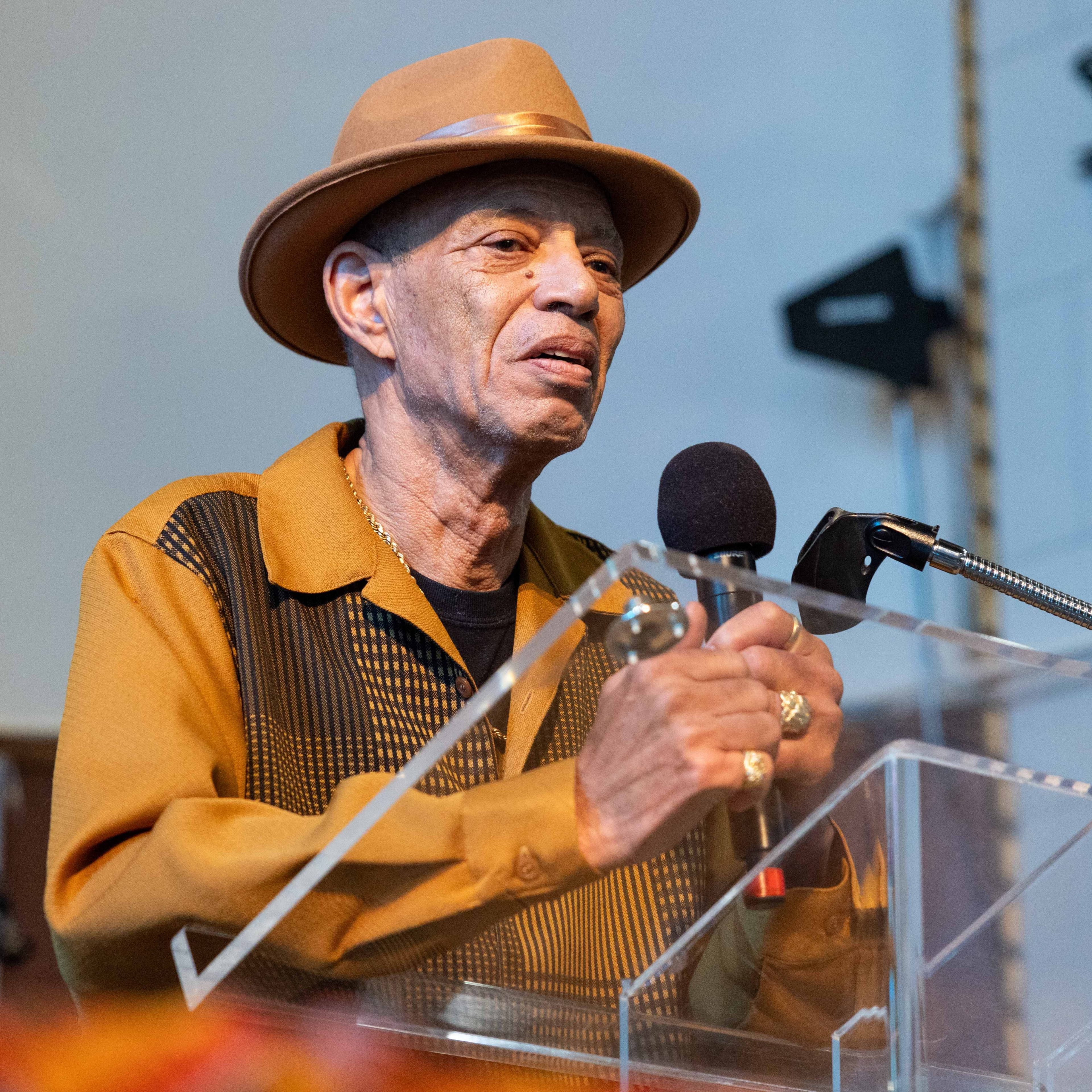 An older man in a brown hat and shirt speaks into a microphone at a podium, with a pensive expression and his hands gesturing slightly.