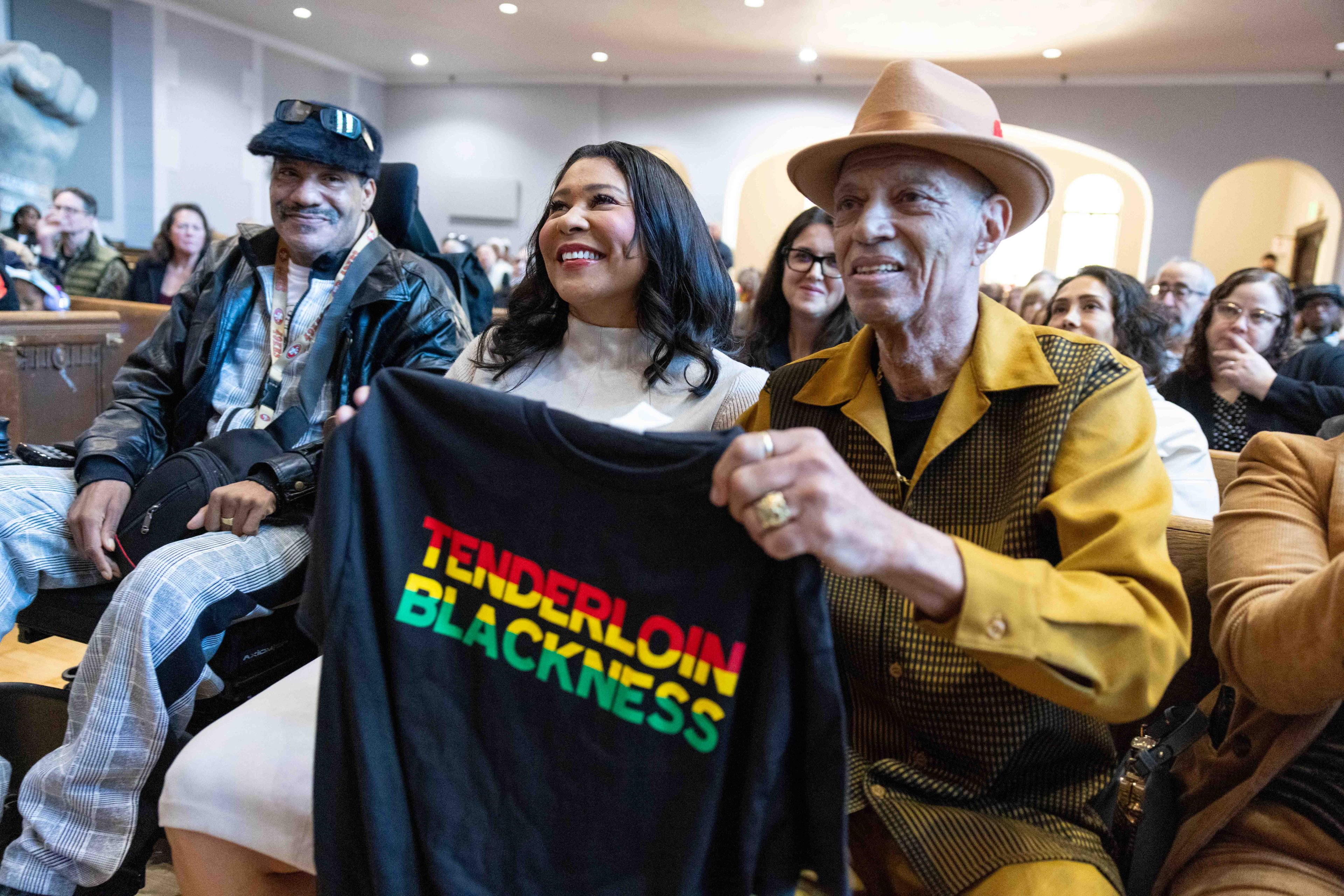 Three people sit together, smiling. The person on the right holds a black shirt with the words &quot;TENDERLOIN BLACKNESS&quot; in red, yellow, and green.