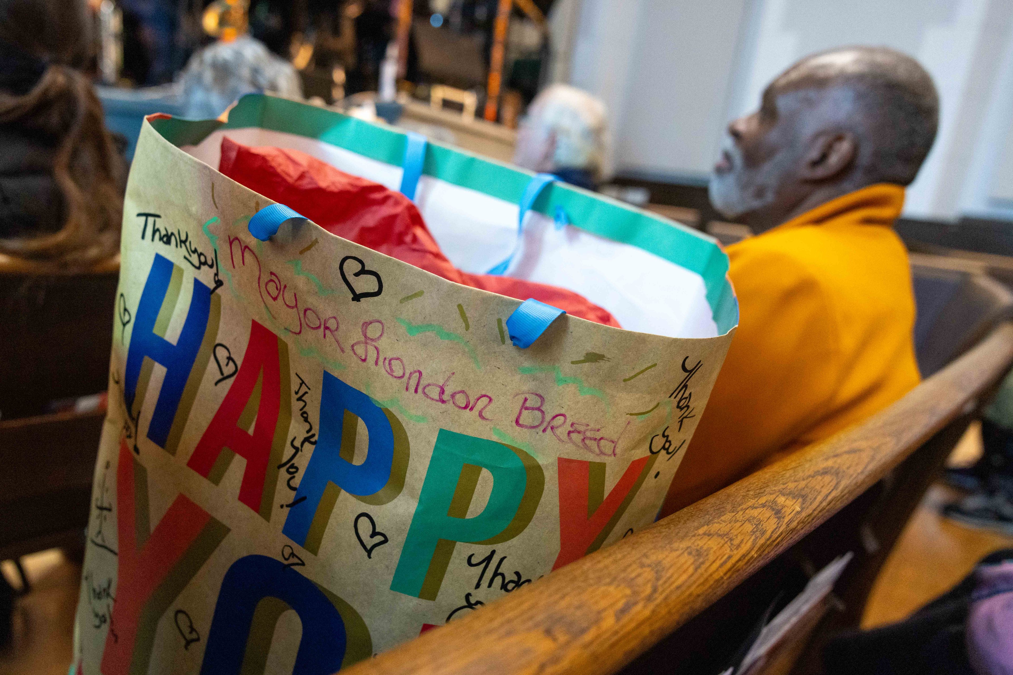 A gift bag with the words &quot;HAPPY&quot; and &quot;Thank you&quot; sits on a wooden bench. Red paper peeks out, and a man in an orange coat is seated nearby in a gathering space.