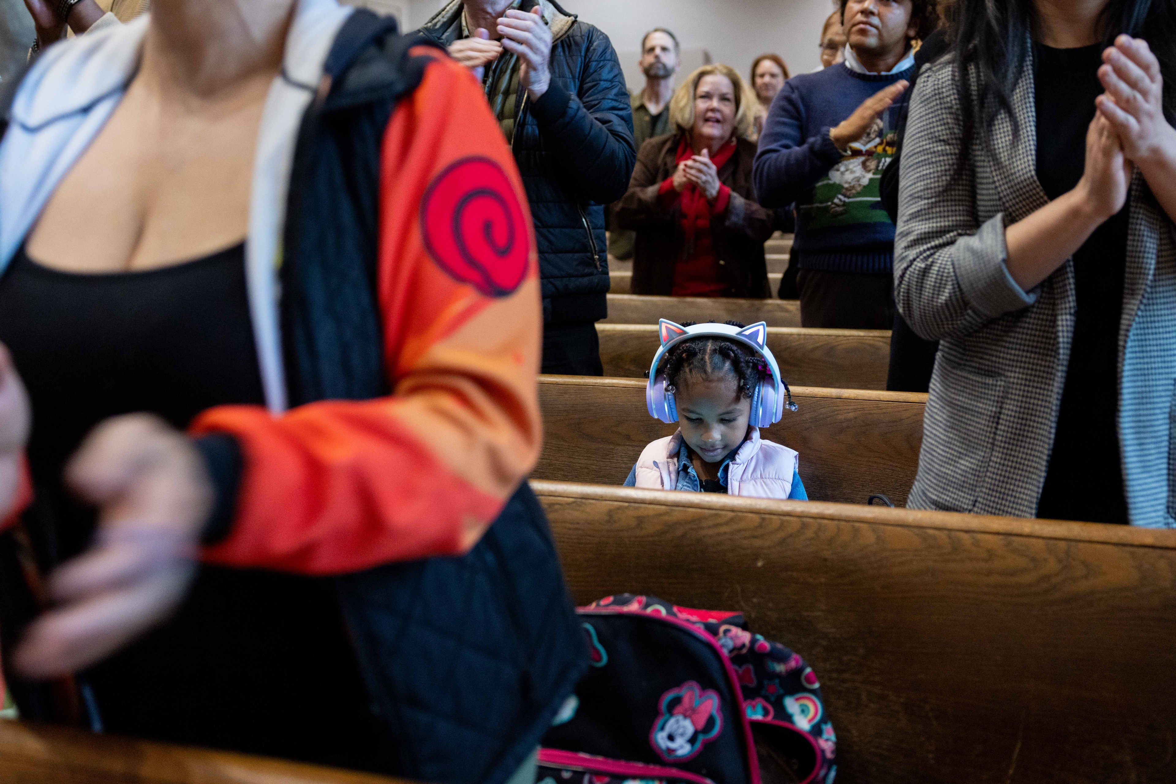 A girl wearing colorful cat-ear headphones sits in a crowded room, focused on her device, amid adults who are standing and clapping.