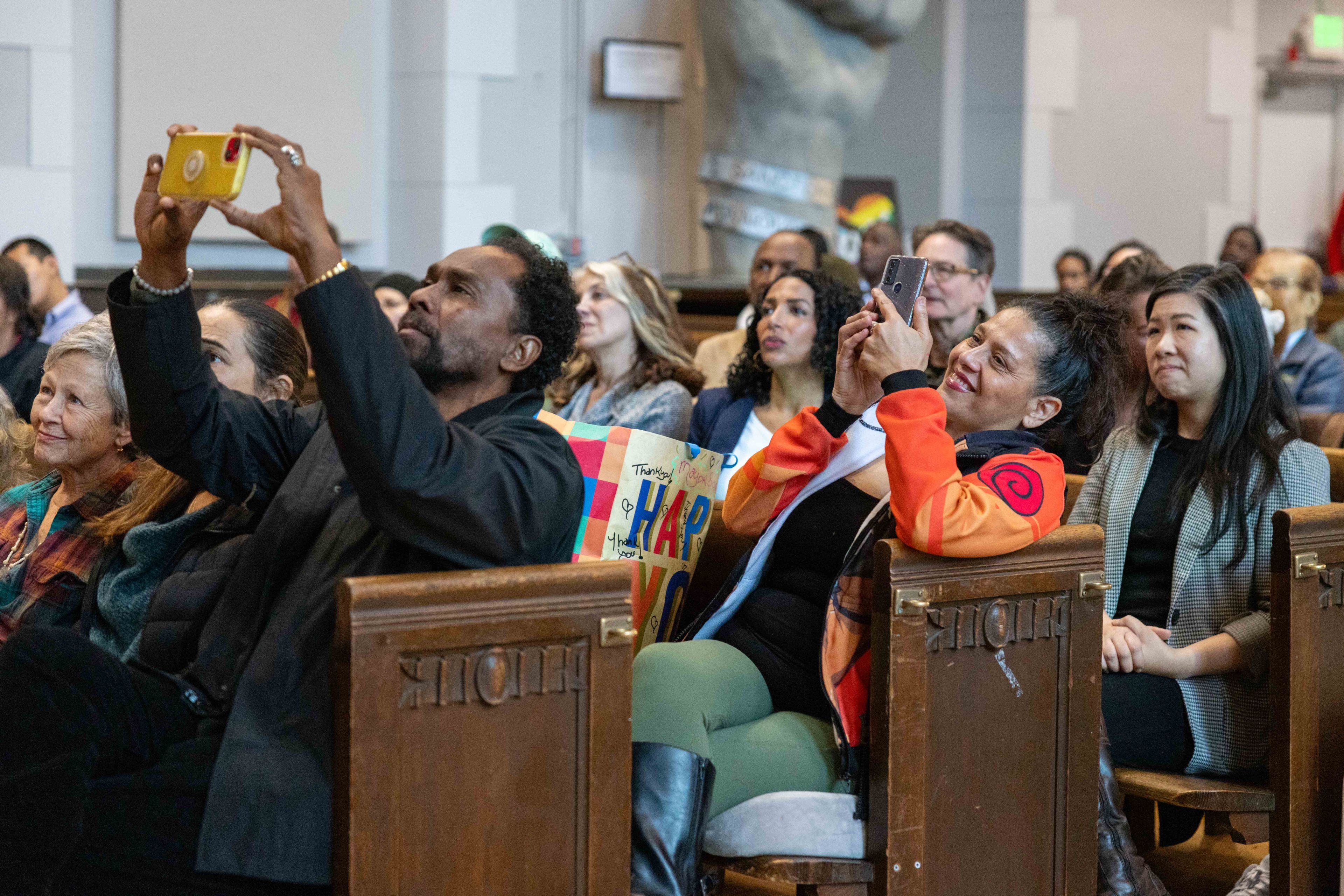 People are seated in what appears to be a hall, with several individuals capturing something on their phones. The atmosphere seems attentive and engaged.