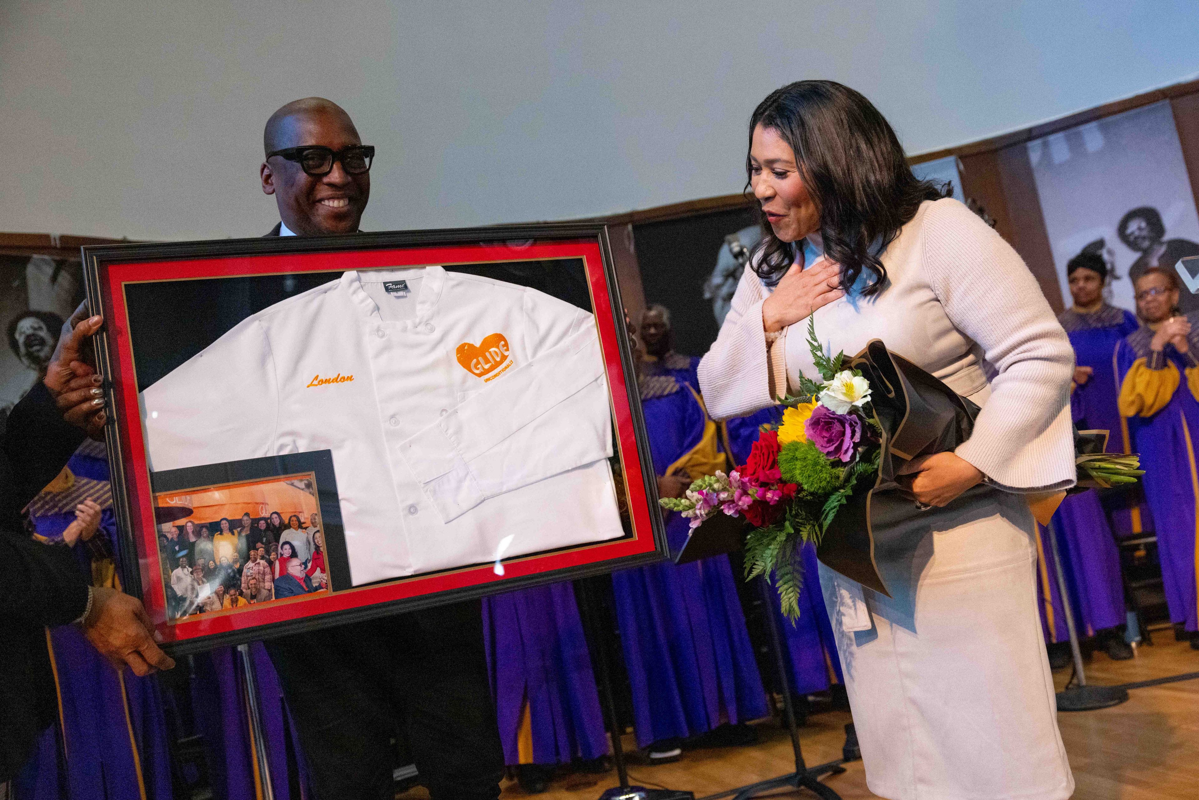 A man presents a framed chef coat and photo to a woman holding a bouquet. She stands with a hand on her chest, surrounded by people in purple and yellow robes.