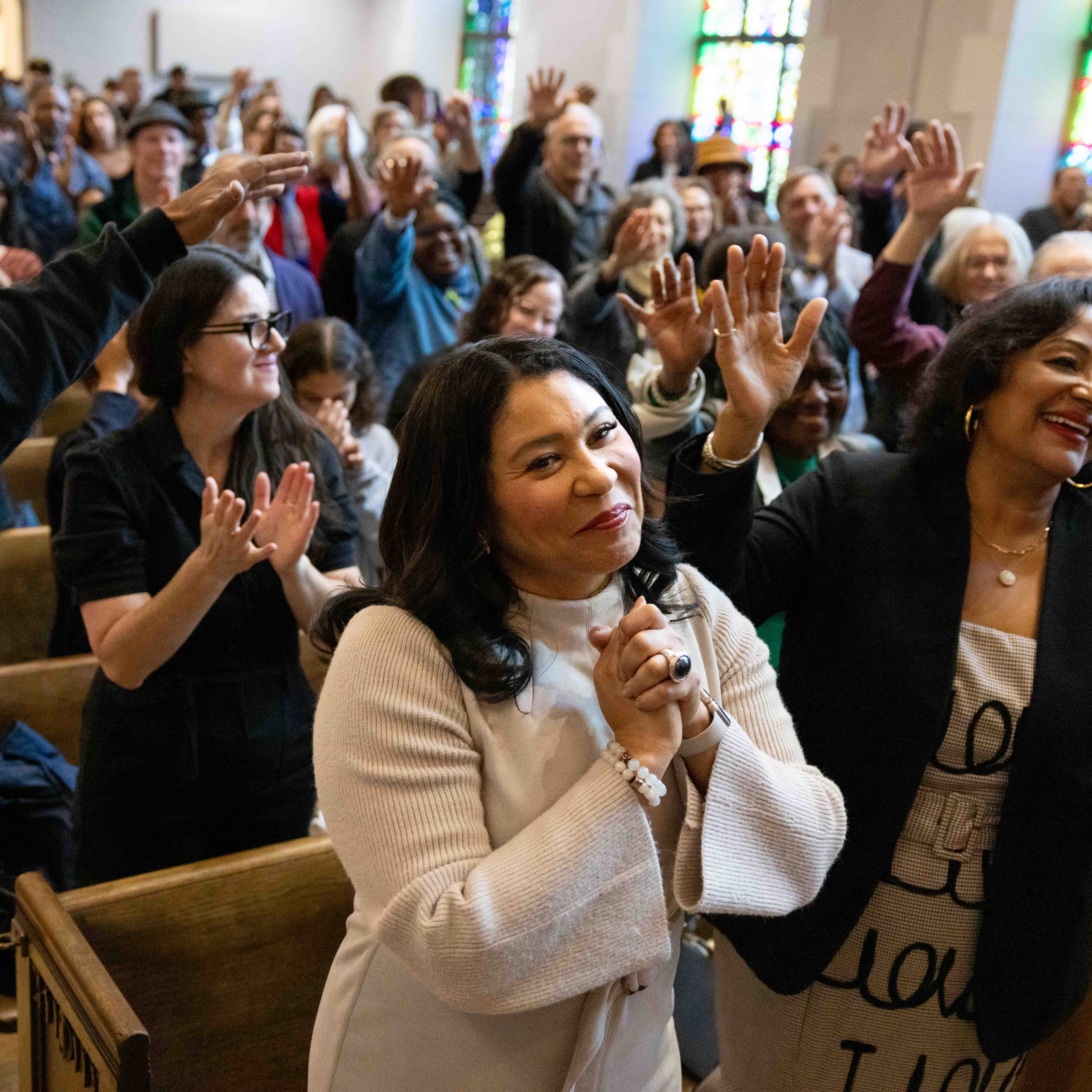 A diverse group of people in a church setting, smiling and clapping with hands raised, surrounded by stained glass windows in the background.