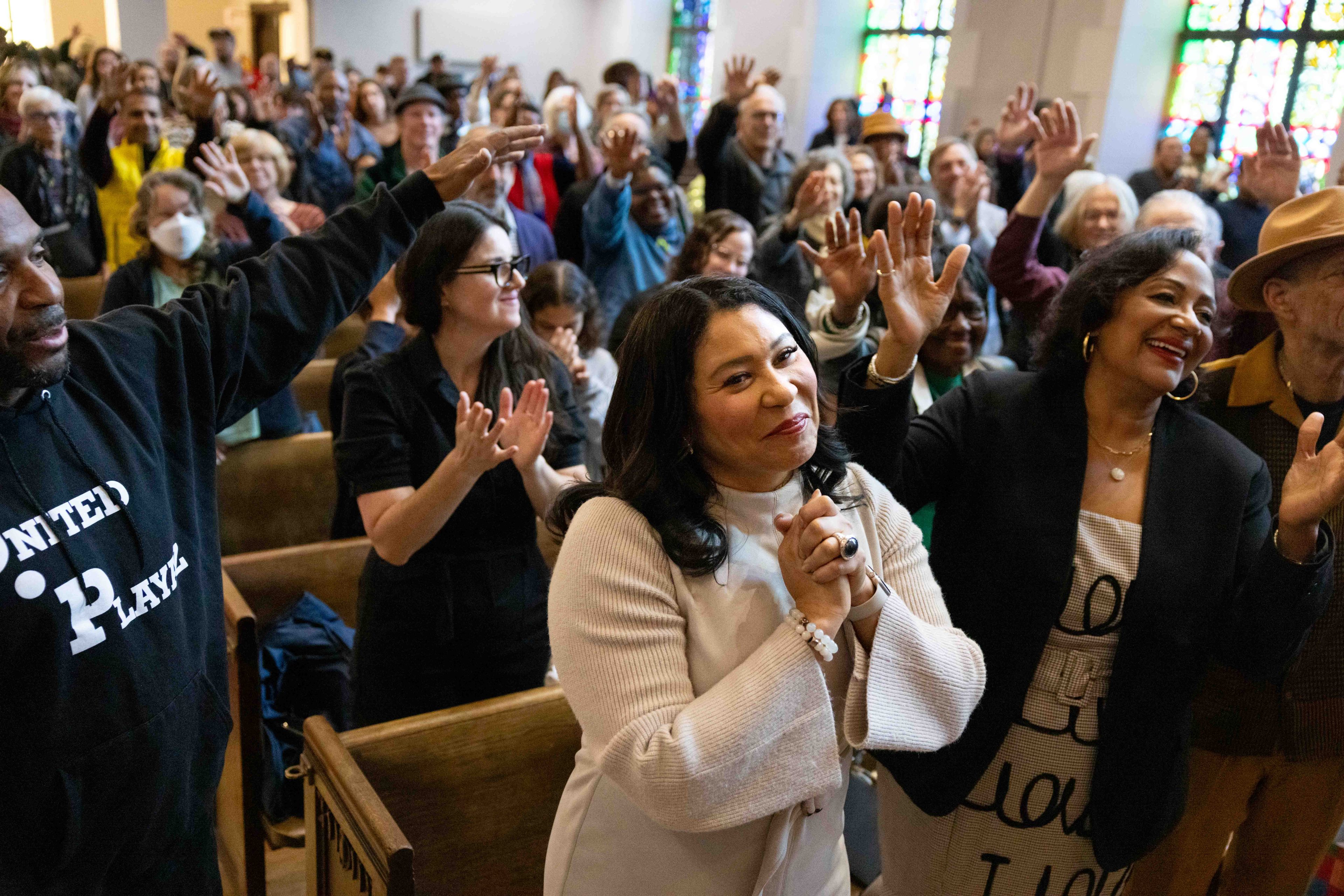 A diverse group of people in a church setting, smiling and clapping with hands raised, surrounded by stained glass windows in the background.