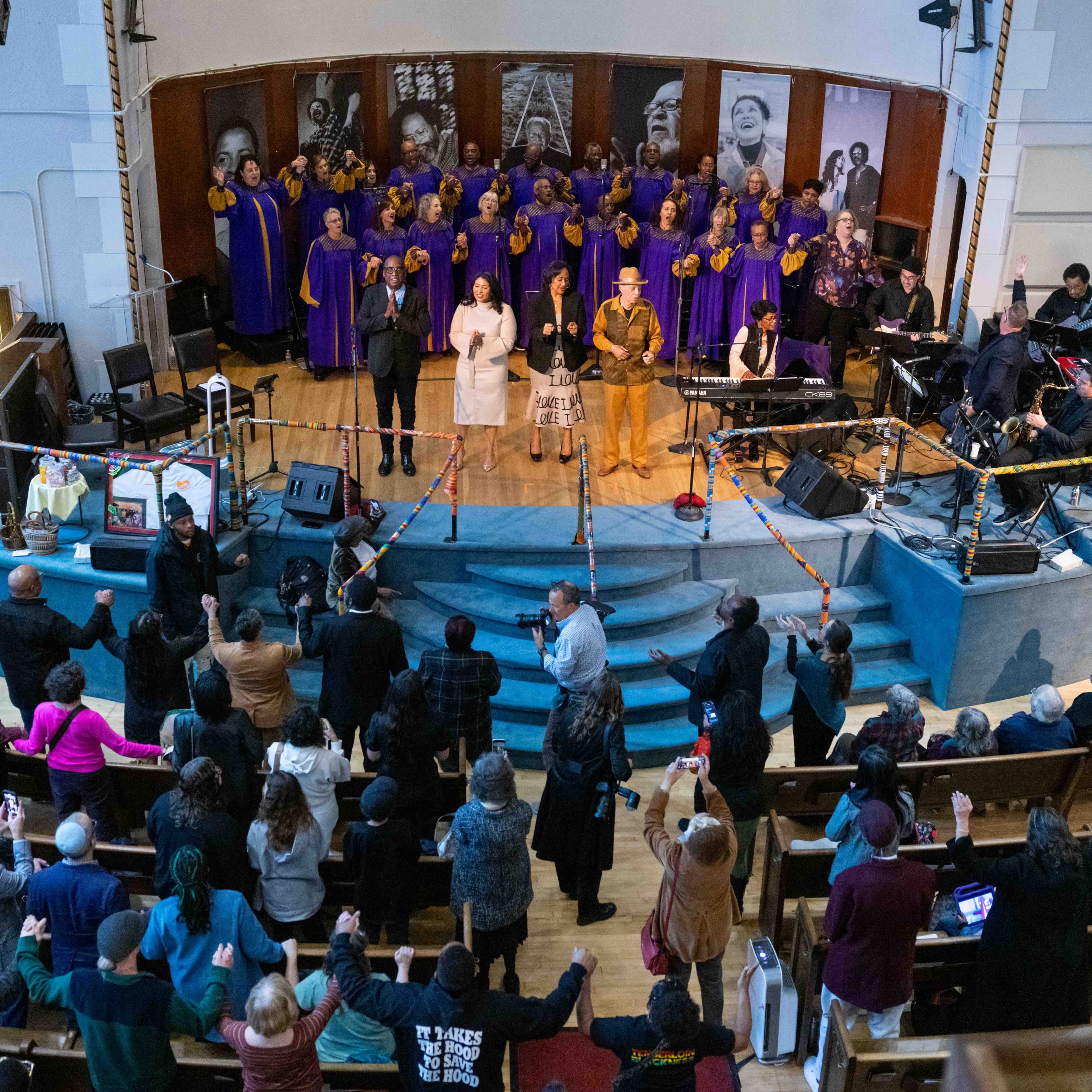 A choir in purple robes performs on stage with musicians, while a diverse audience stands, some holding hands, in a warmly lit church setting.