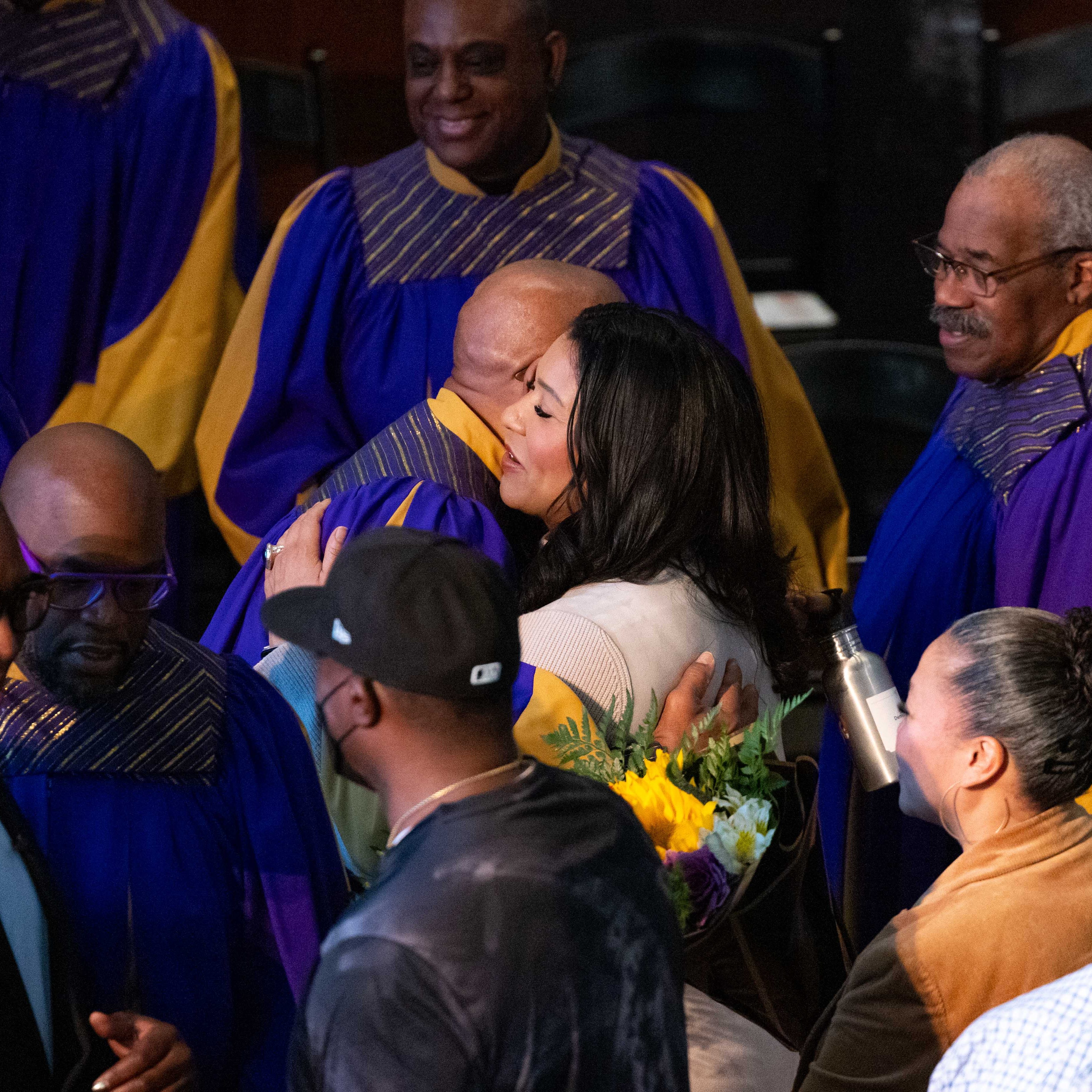 A woman hugs a choir member in a purple and yellow robe, surrounded by other people and choir members. She holds a bouquet of yellow flowers.