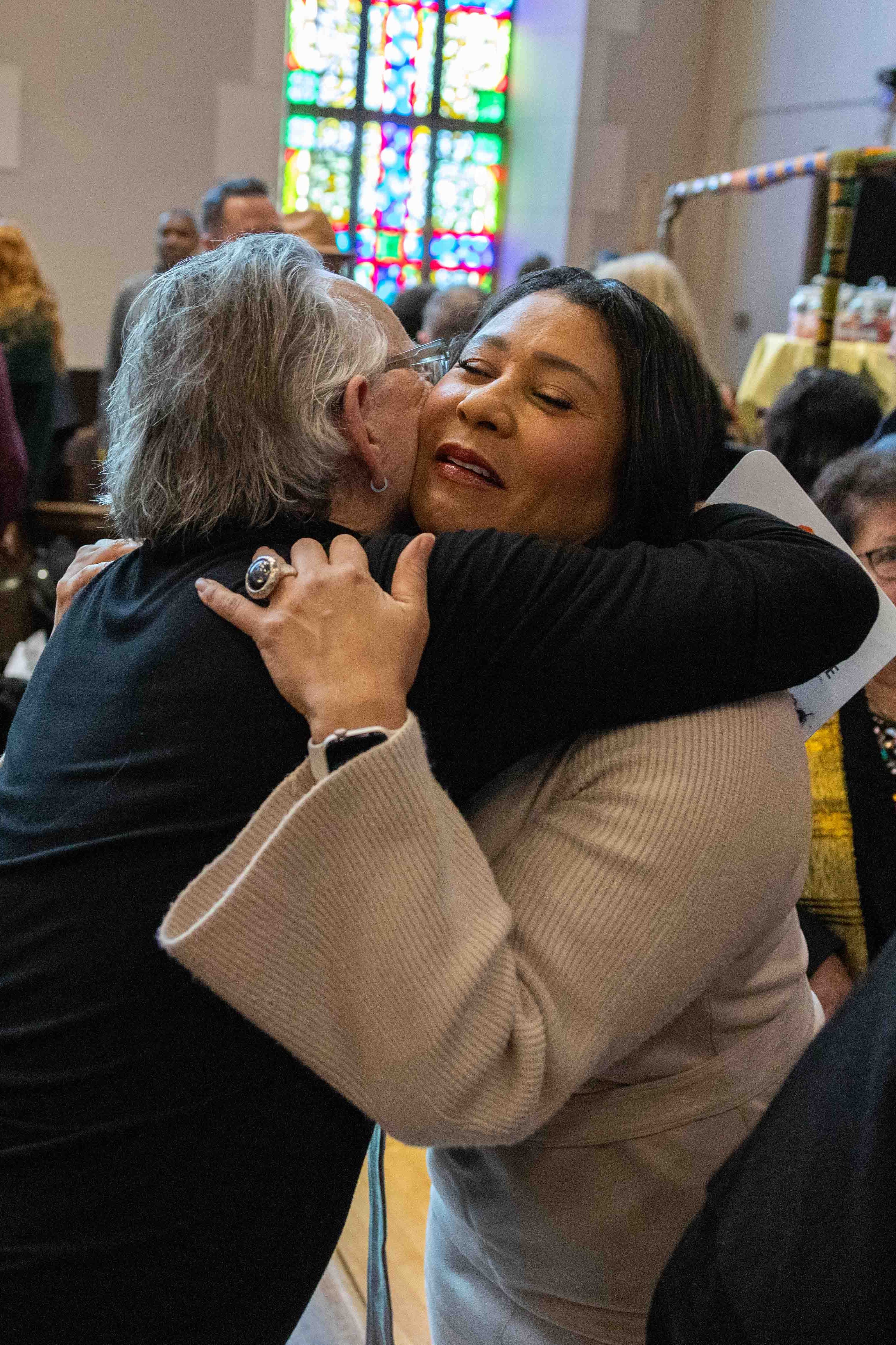 Two people embrace in a warm hug inside a gathering space with stained glass windows. Other individuals are around, creating a lively, friendly atmosphere.