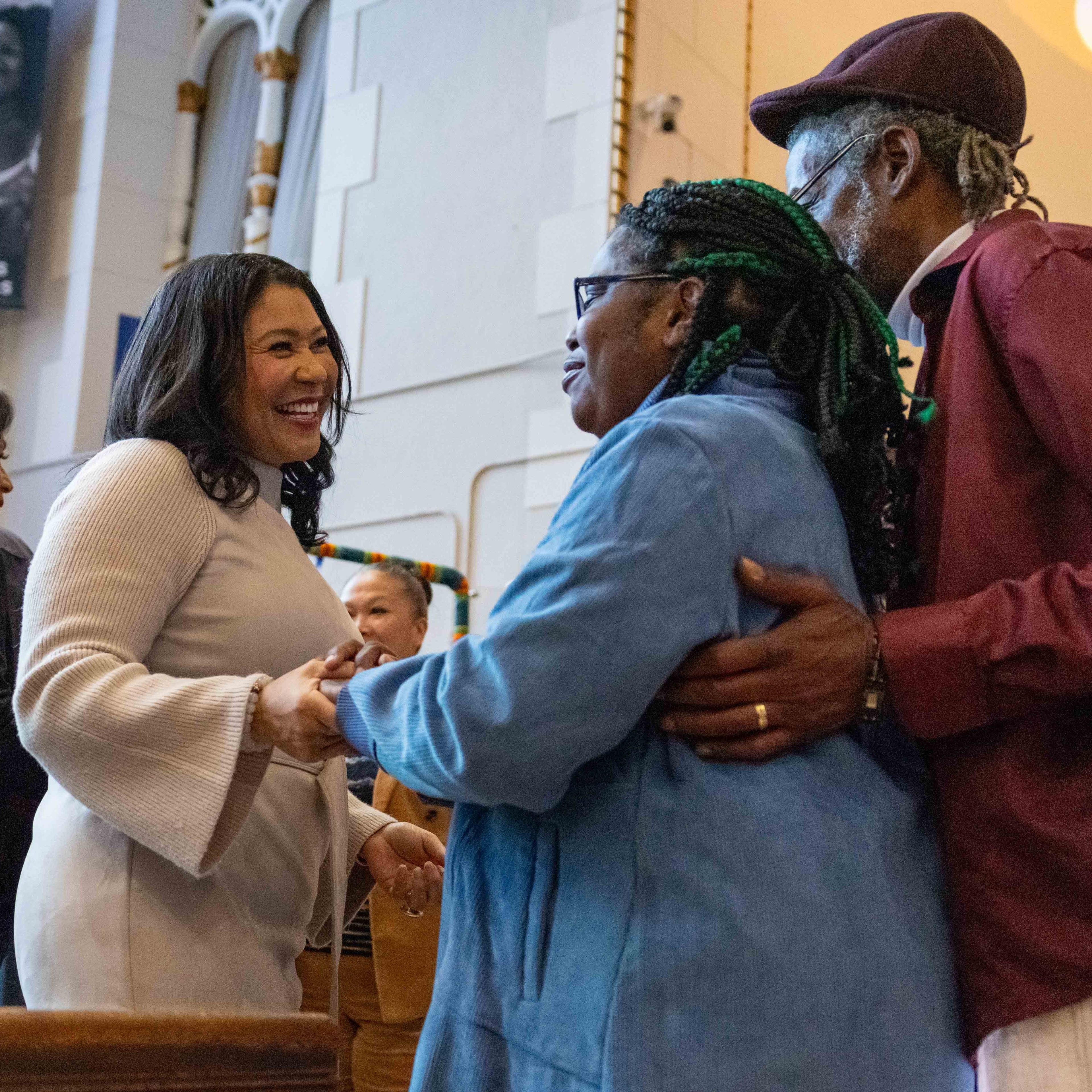 A smiling woman in a light sweater engages warmly with a couple, surrounded by people in an ornate, colorful setting with stained glass and banners.