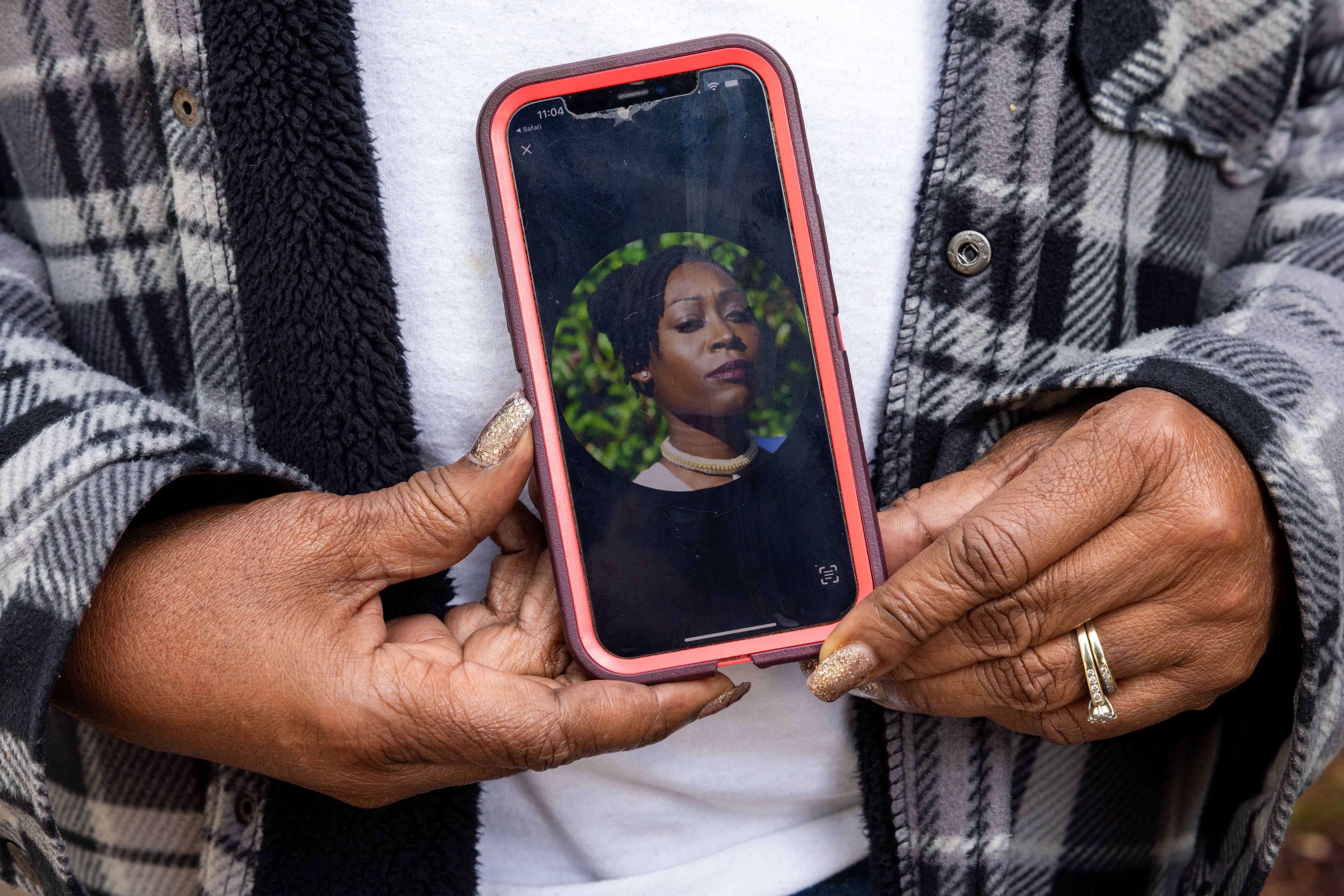 A pair of hands with glittery nails and a ring hold a smartphone showing a woman's photo. She's wearing a necklace and standing against a green background.