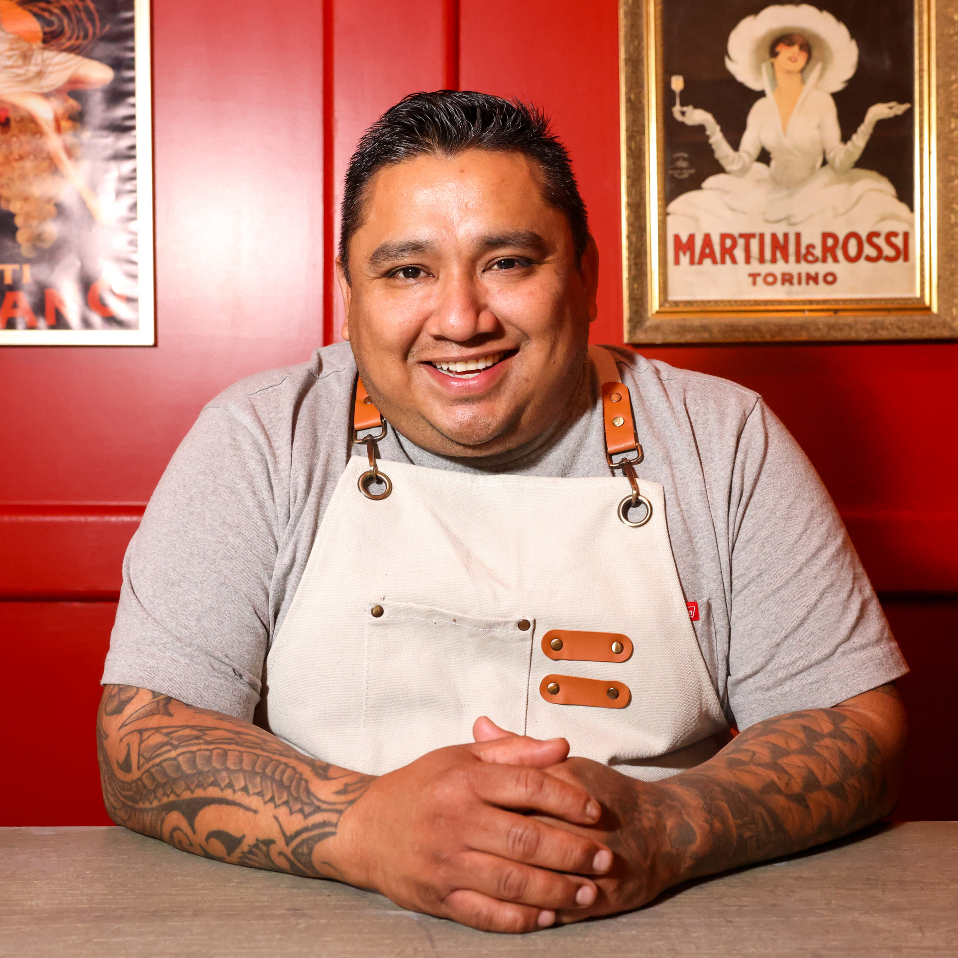 A smiling person with tattooed arms sits at a table, wearing a gray shirt and a white apron against a red background with vintage posters.