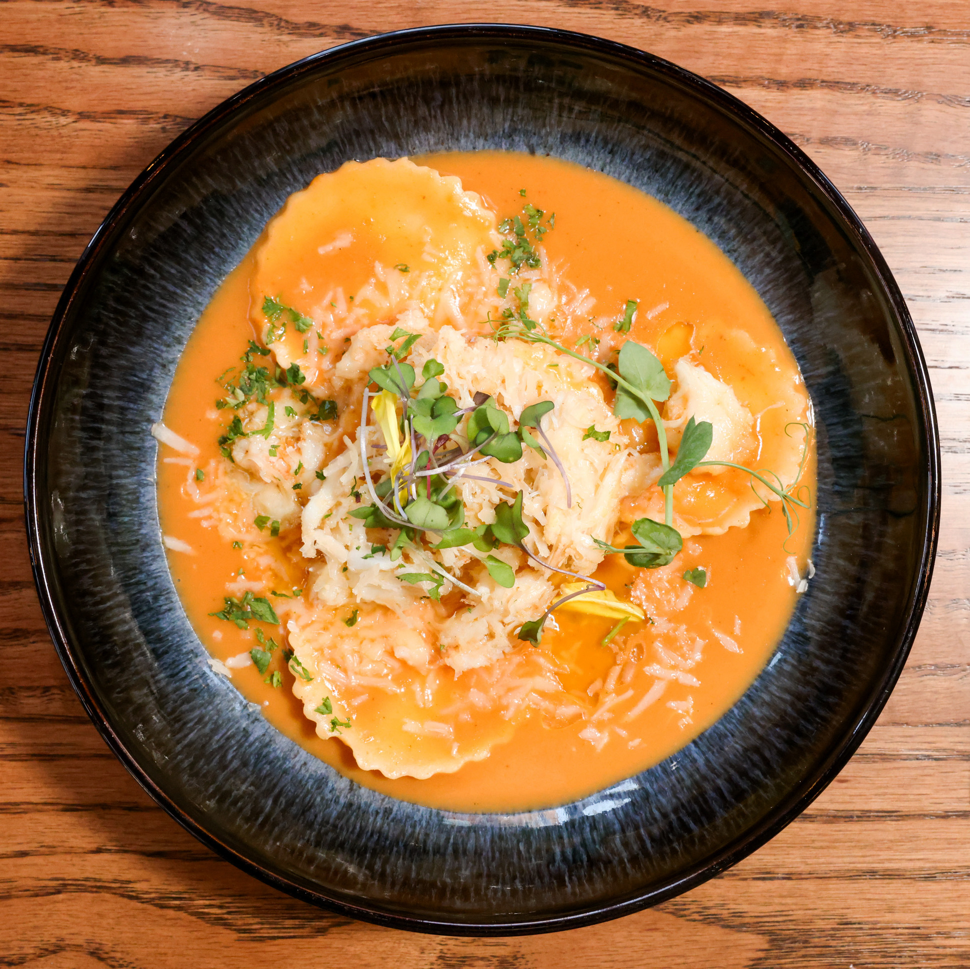 A black bowl holds ravioli in a creamy orange sauce, topped with grated cheese and fresh green microgreens, placed on a wooden table.