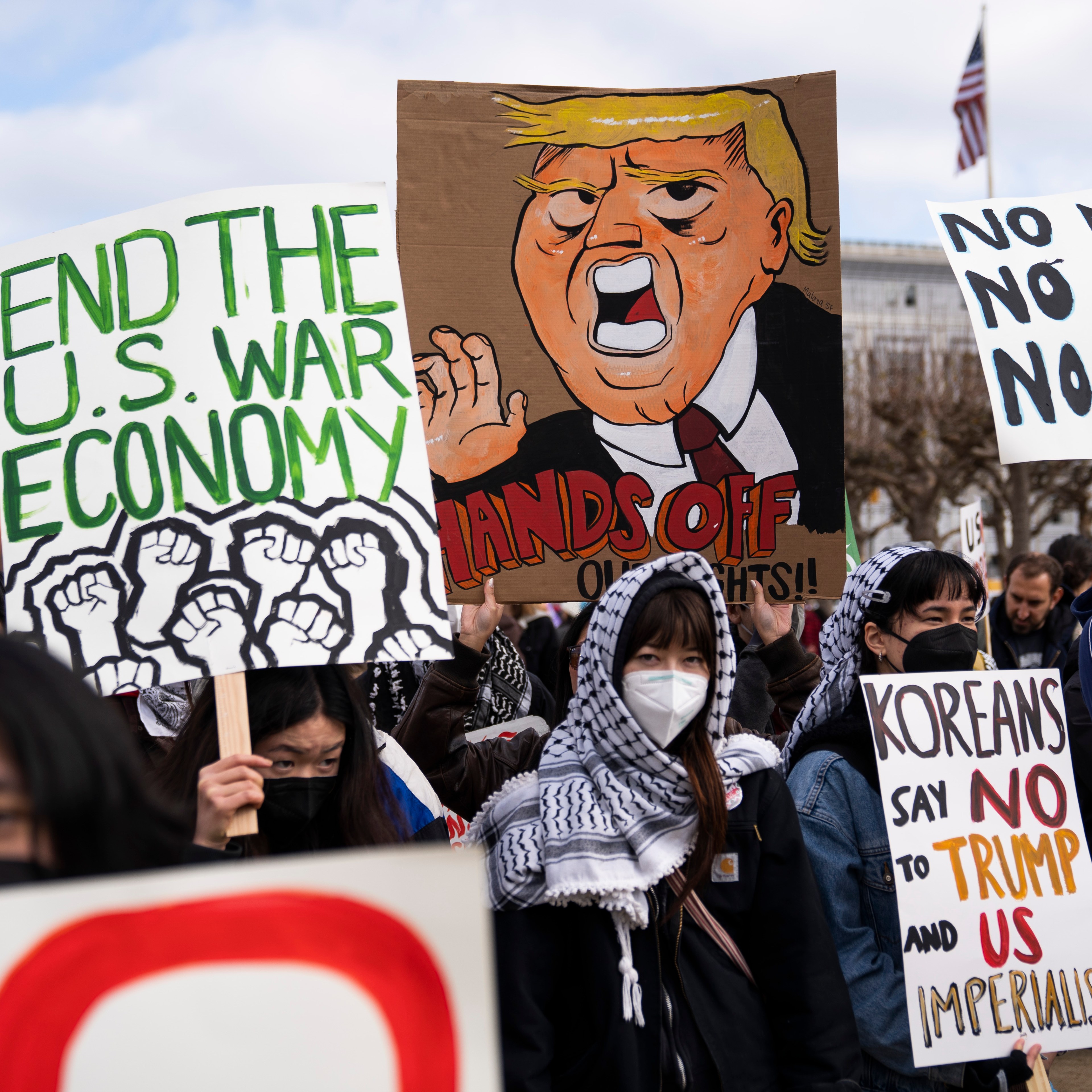 A group of protesters holds signs with messages opposing war, Trump, and imperialism. One sign features a caricature, while others emphasize ending the U.S. war economy.