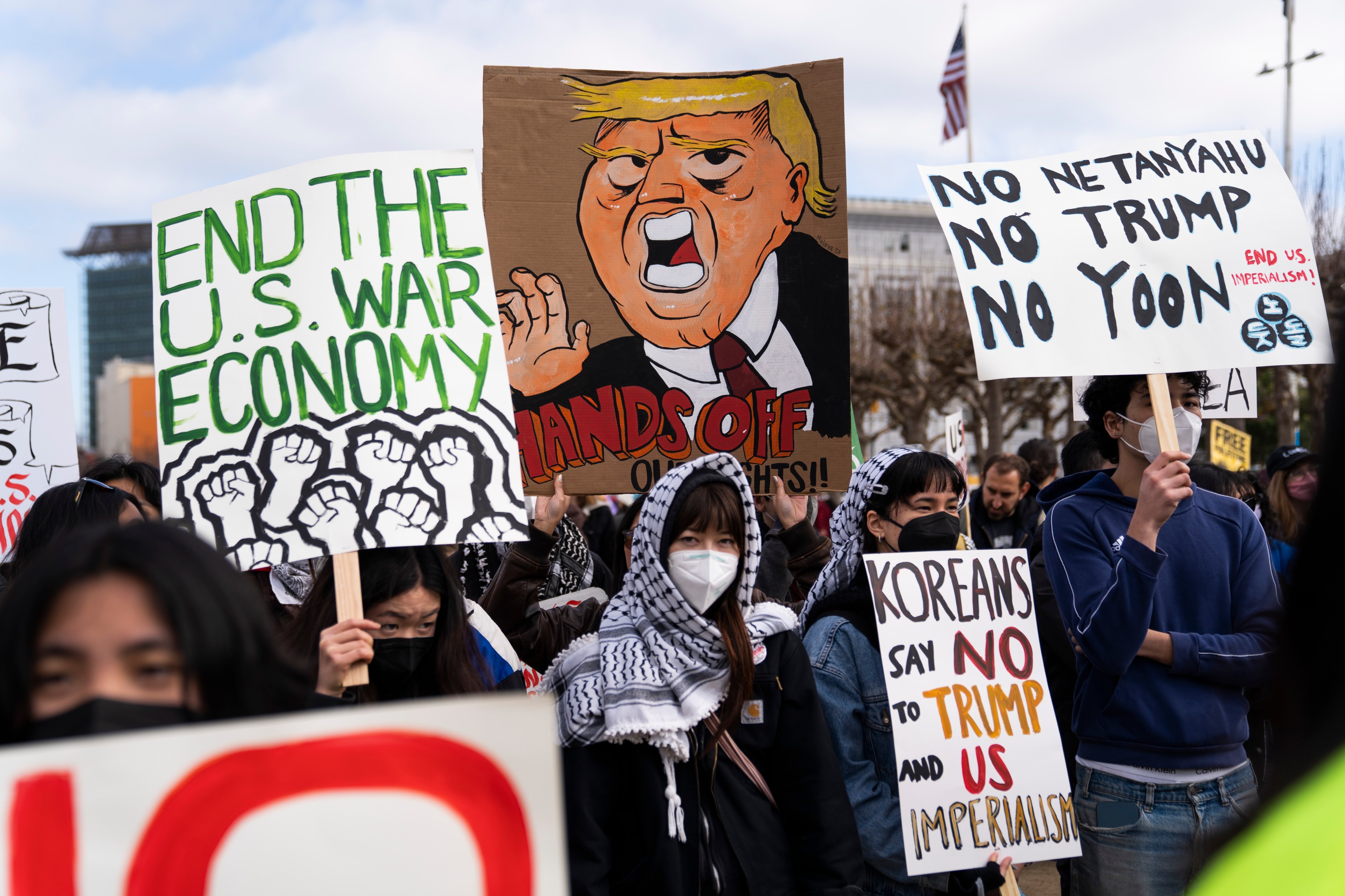 A group of protesters holds signs with messages opposing war, Trump, and imperialism. One sign features a caricature, while others emphasize ending the U.S. war economy.