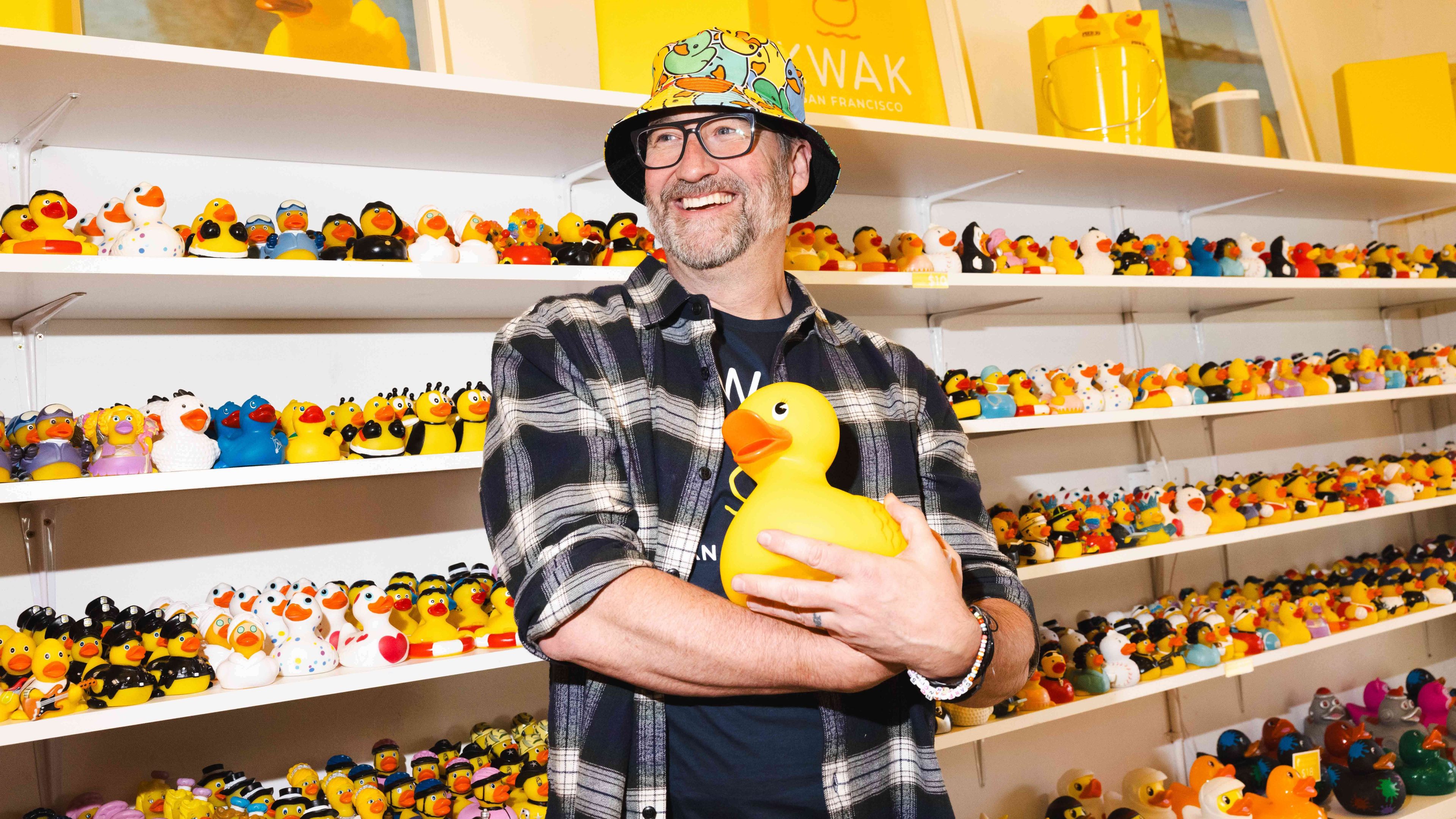 A person in a checkered shirt and a duck-themed hat is smiling, holding a large yellow rubber duck, surrounded by shelves filled with various rubber ducks.