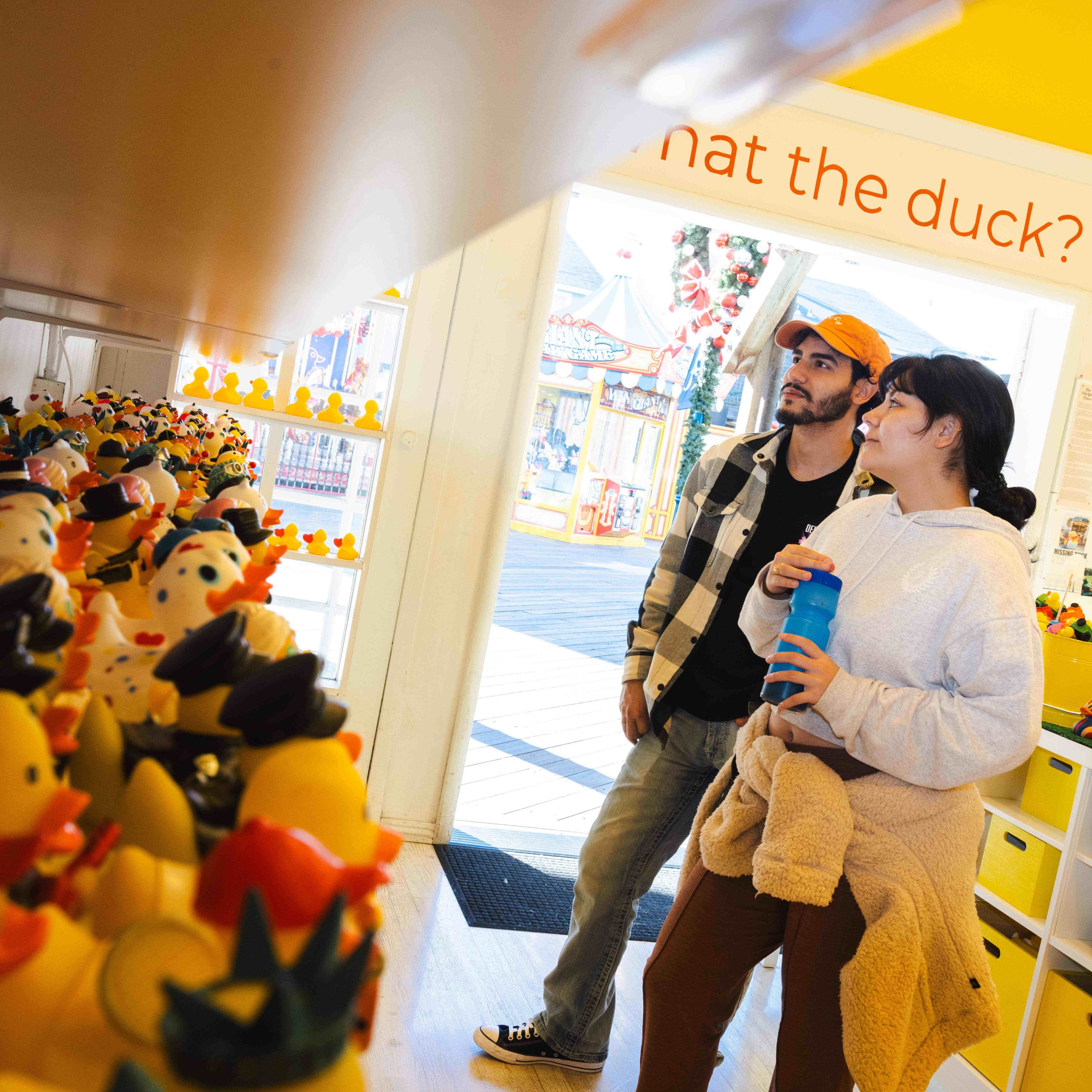 A man and woman look at rows of themed rubber ducks in a brightly lit shop. A sign above reads, &quot;What the duck?&quot; They're casually dressed, and the scene feels playful.