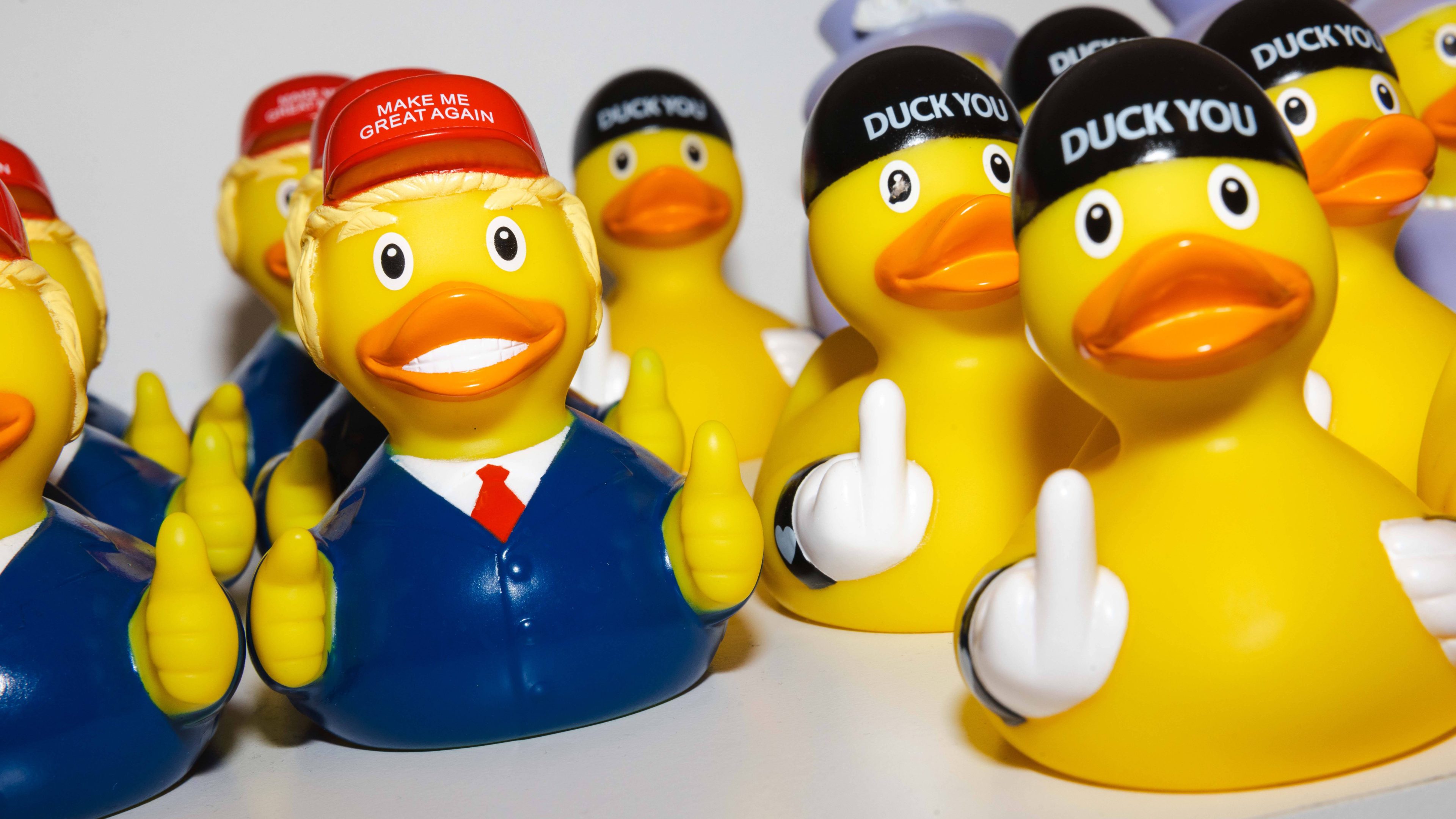 The image shows yellow rubber ducks. Some wear red or black hats with bold text. They're posed with outstretched wings mimicking a playful rude gesture.