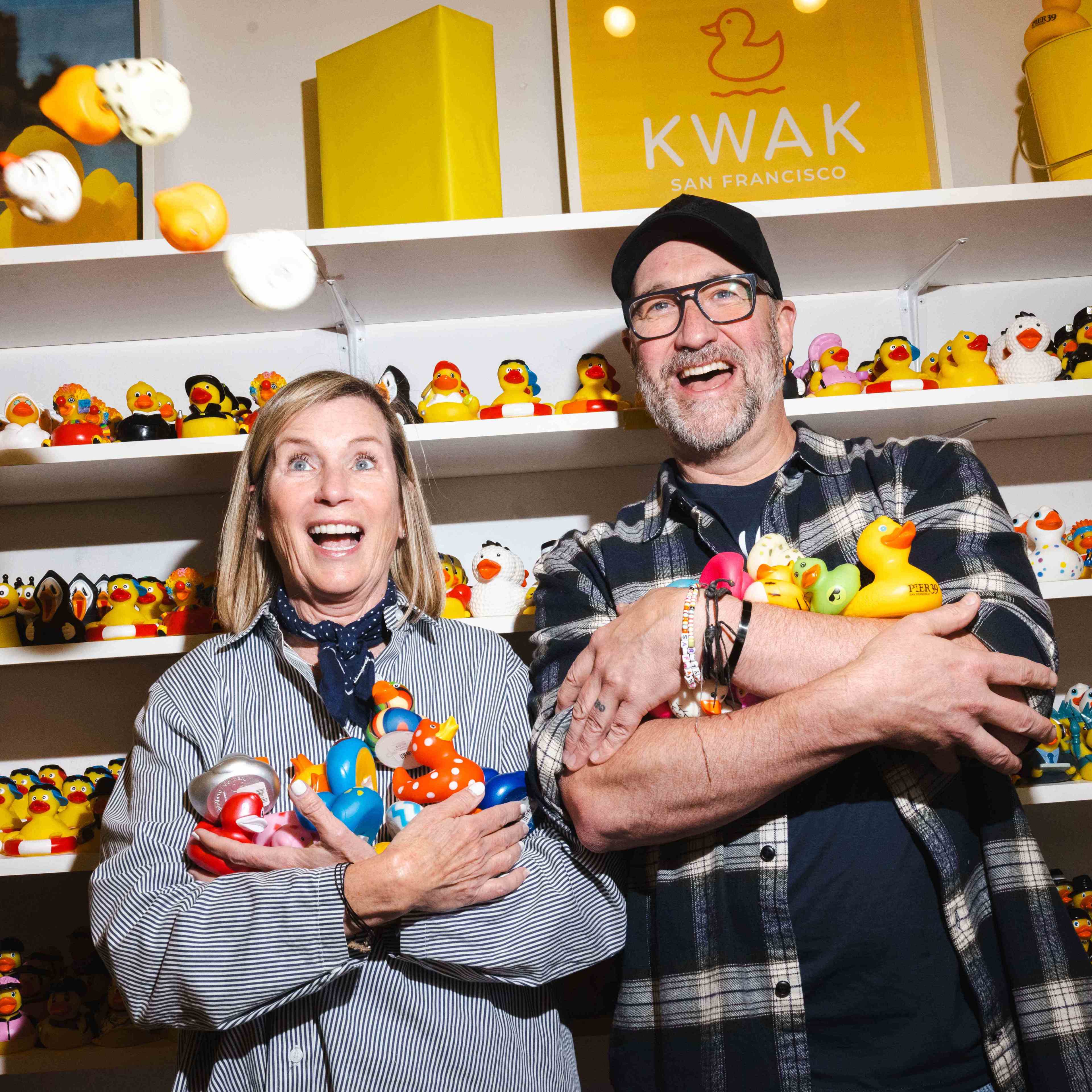 A smiling woman and man stand in front of shelves filled with rubber ducks, each holding several ducks in their arms. Bright lighting highlights their joyful expressions.