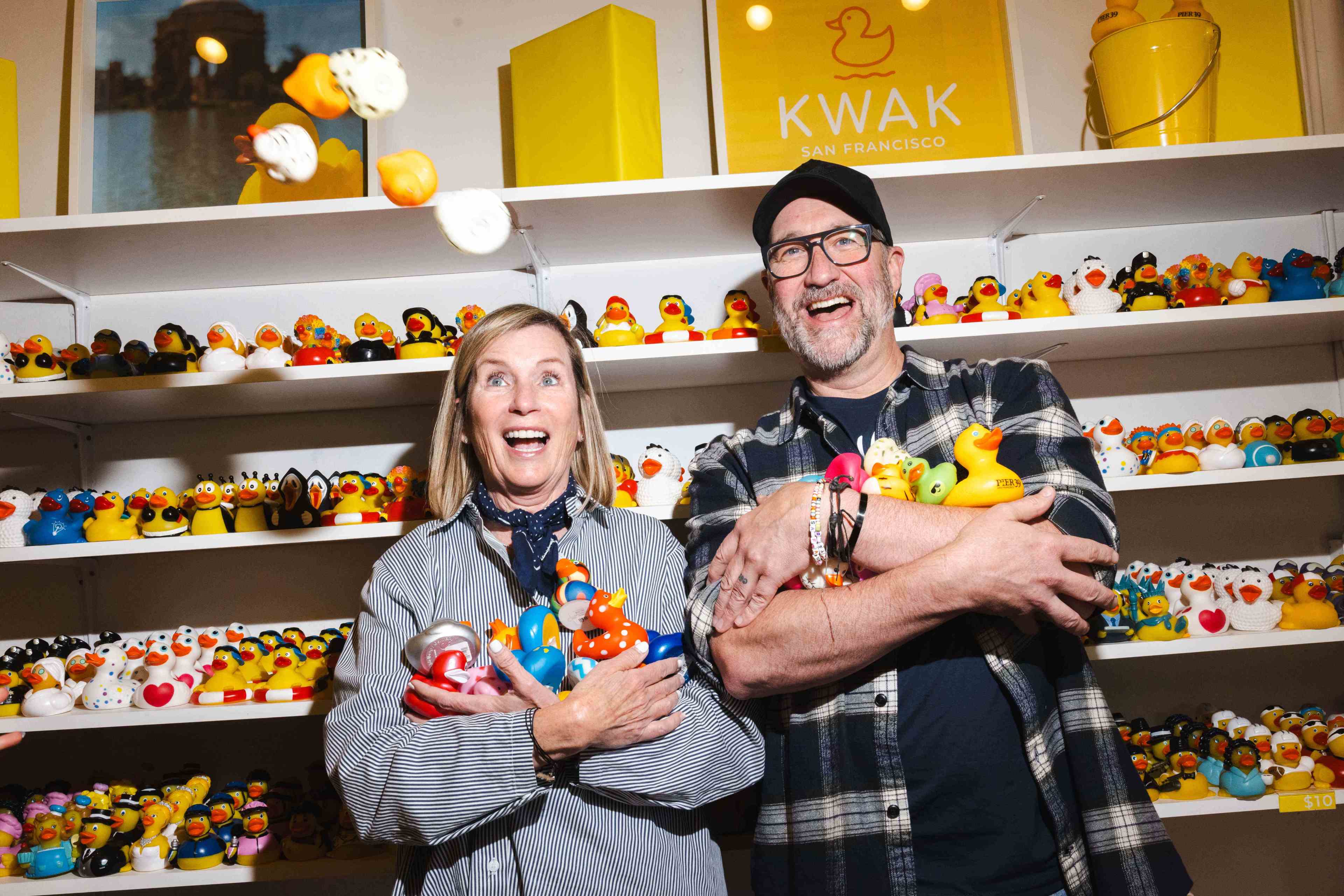 A smiling woman and man stand in front of shelves filled with rubber ducks, each holding several ducks in their arms. Bright lighting highlights their joyful expressions.