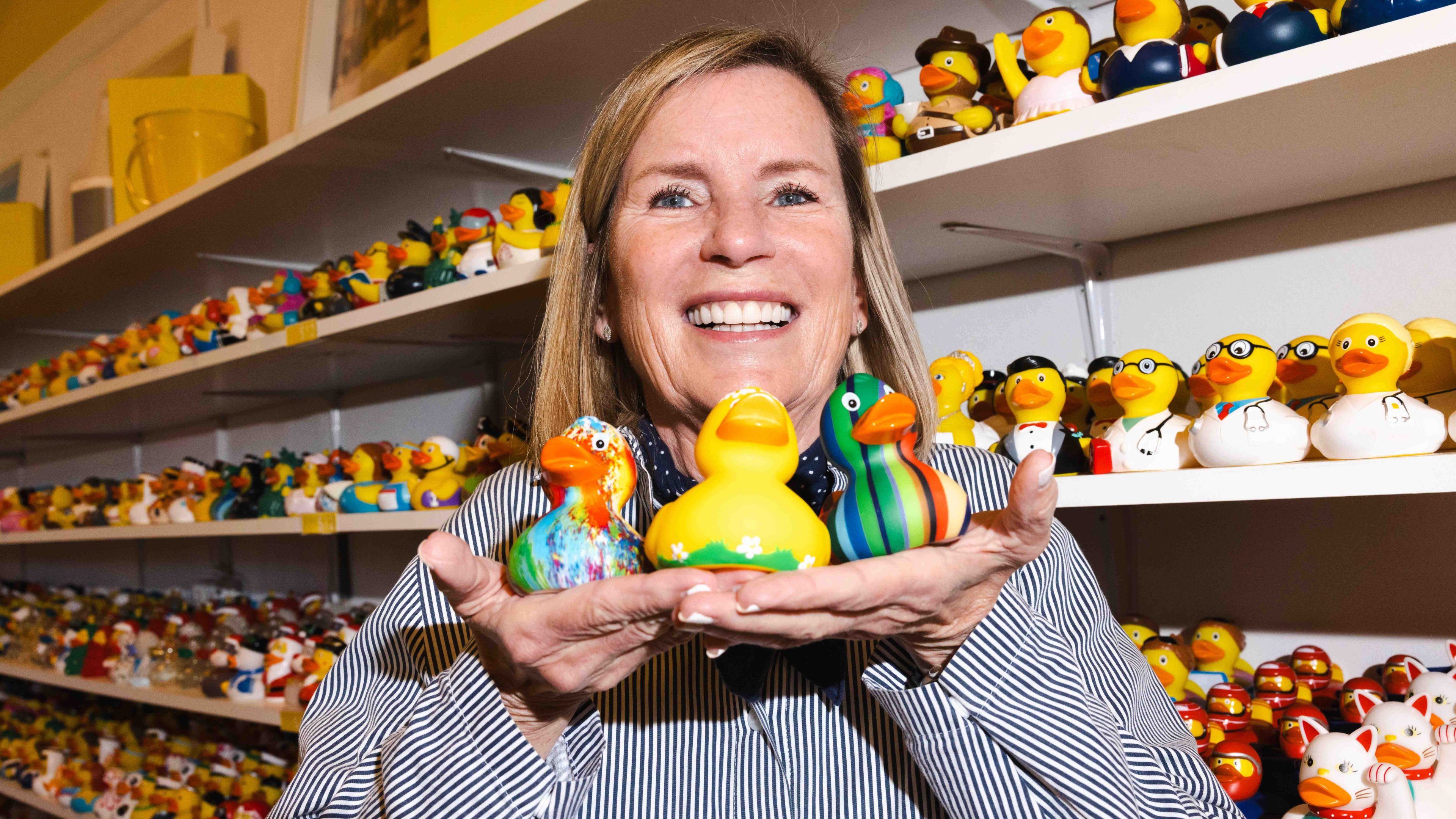 A smiling woman holds four colorful rubber ducks, standing in front of shelves filled with various other rubber ducks.