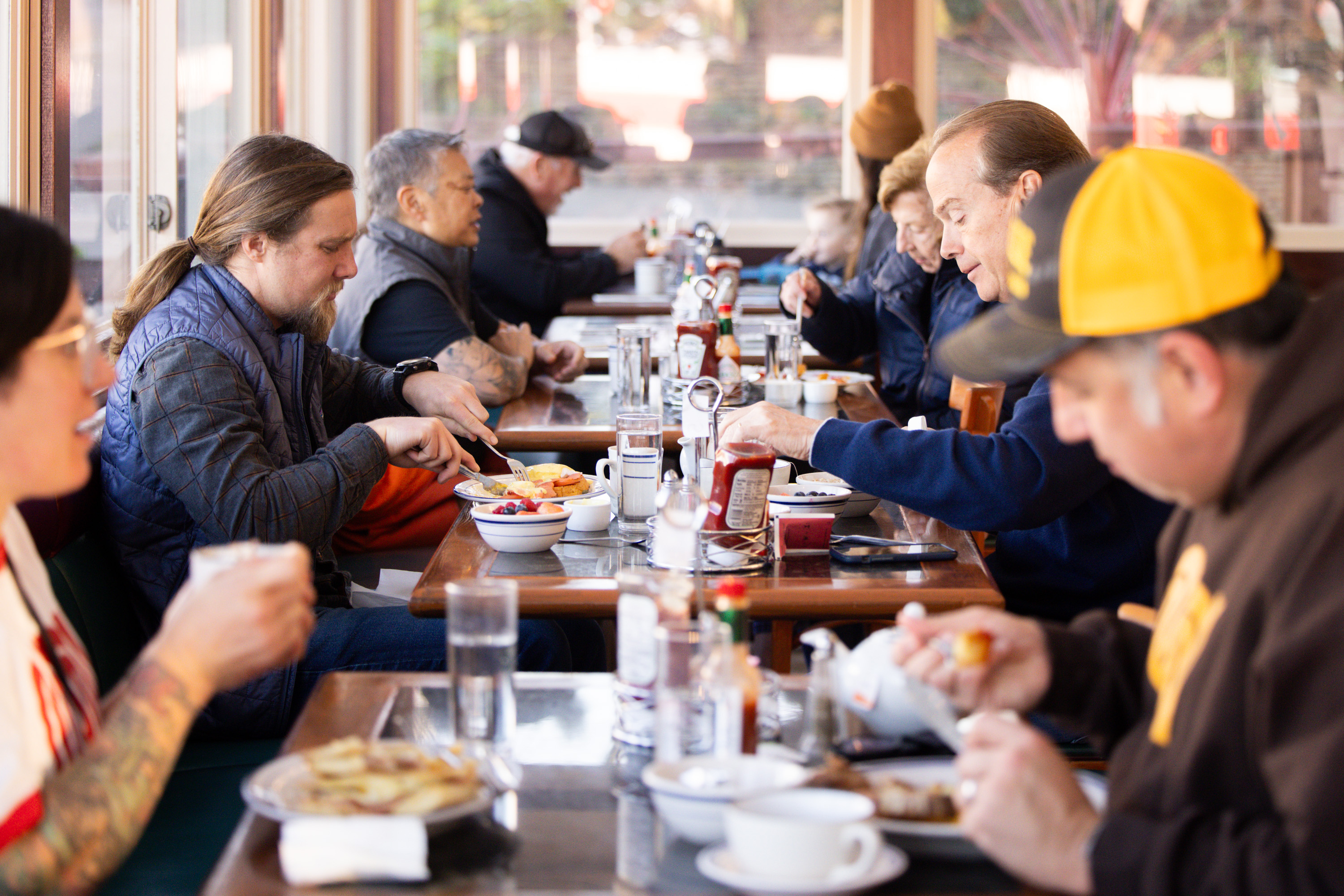 People are seated at a long table in a diner, eating breakfast. They have plates of food, bowls of fruit, and beverages, with condiments and utensils on the table.