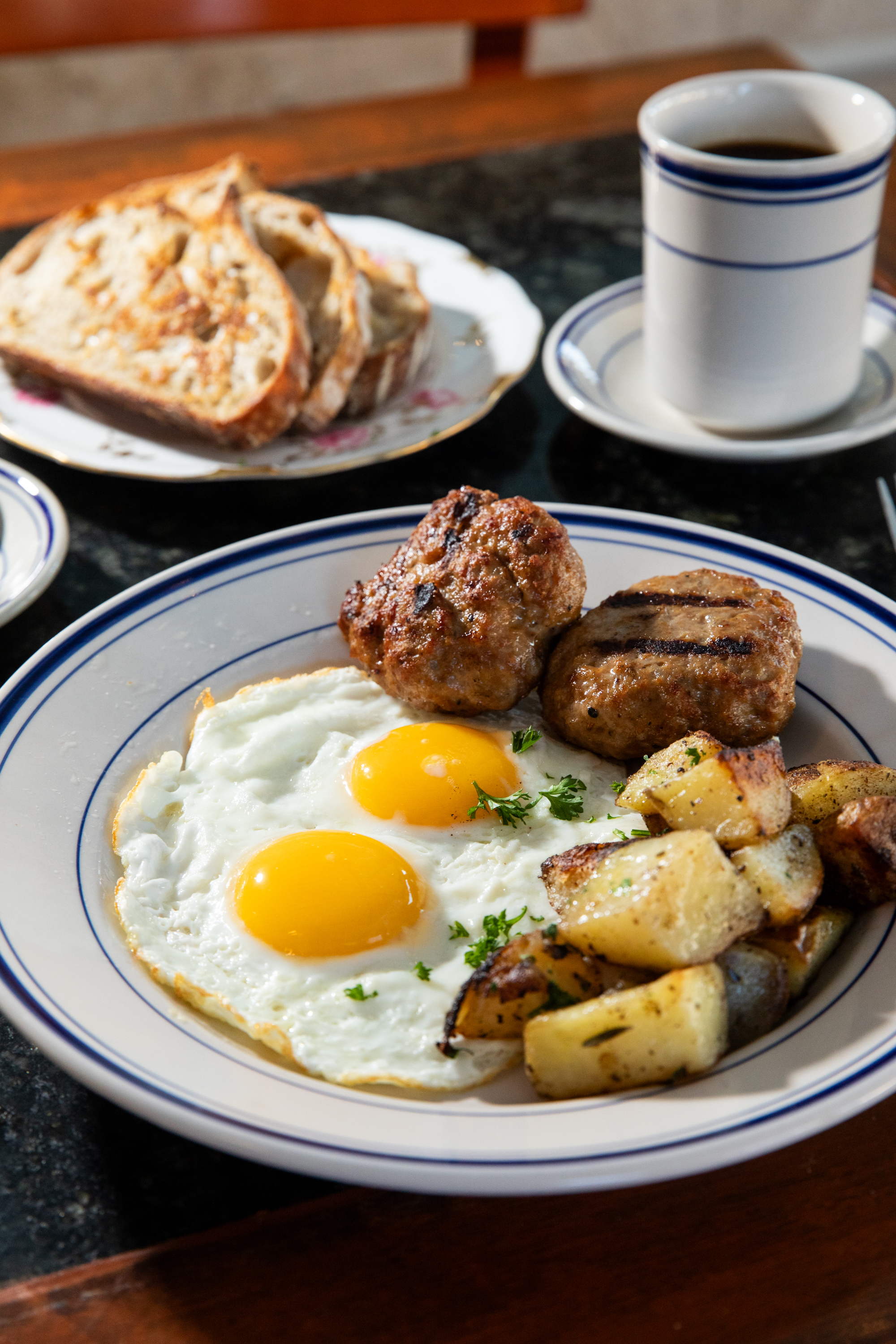 A hearty breakfast featuring two sunny-side-up eggs, sausage, and seasoned potatoes on a plate. Toast and a cup of black coffee are set beside it.