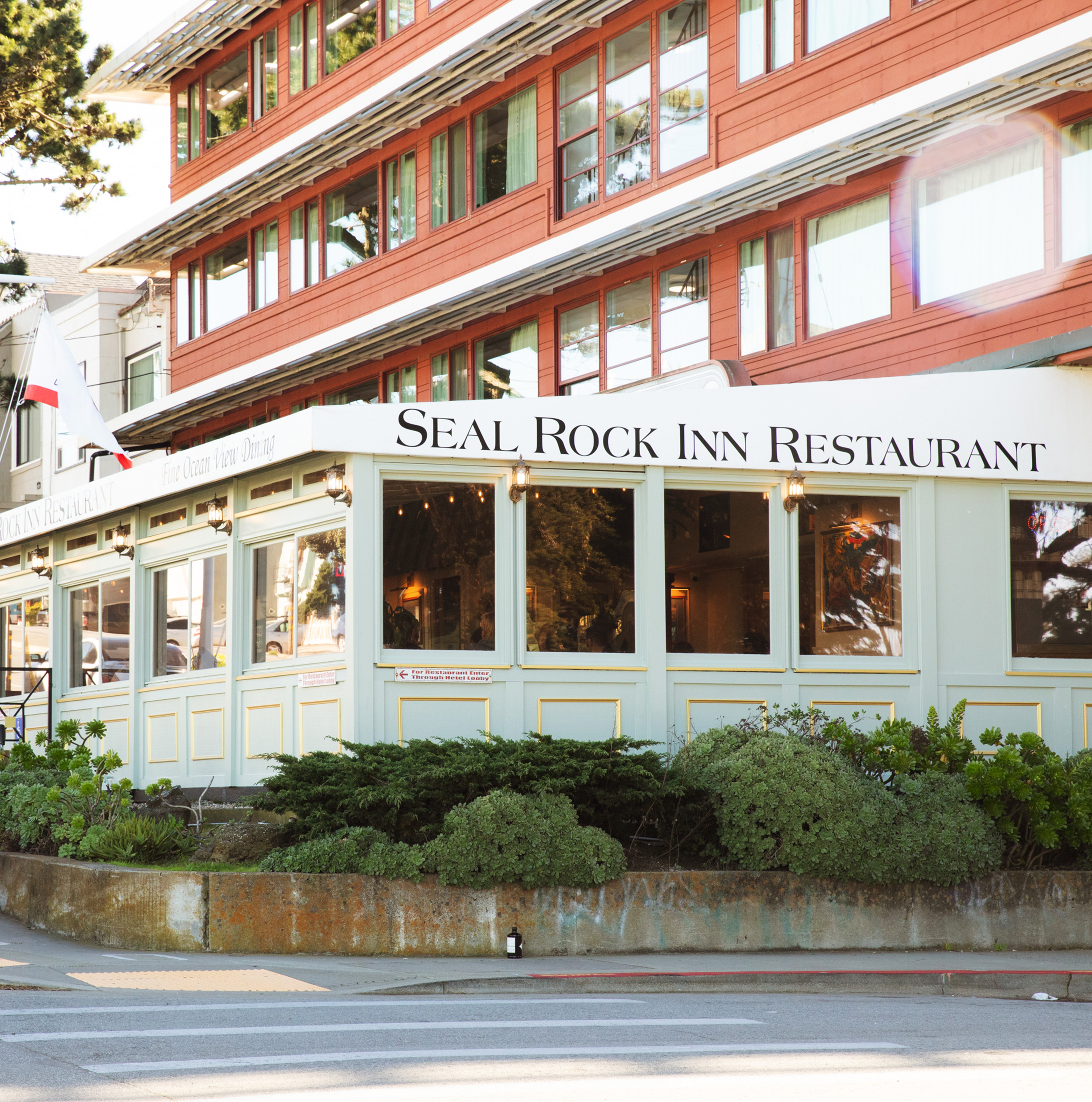 The image shows a restaurant with a sign reading &quot;Seal Rock Inn Restaurant.&quot; It has large windows, a green exterior, and surrounding greenery.