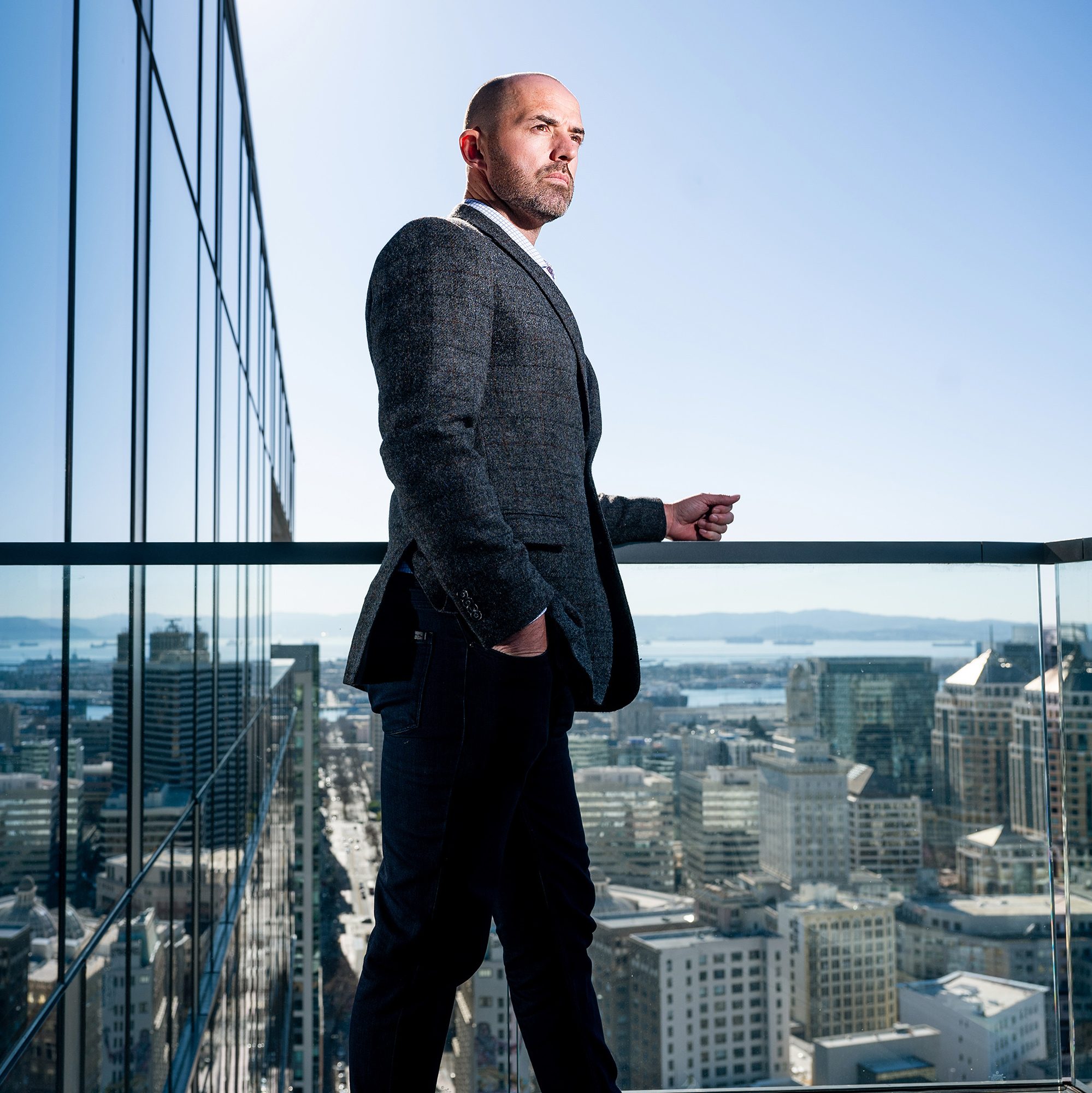 A man in a suit stands on a balcony overlooking a cityscape, with glass buildings and a clear blue sky in the background.