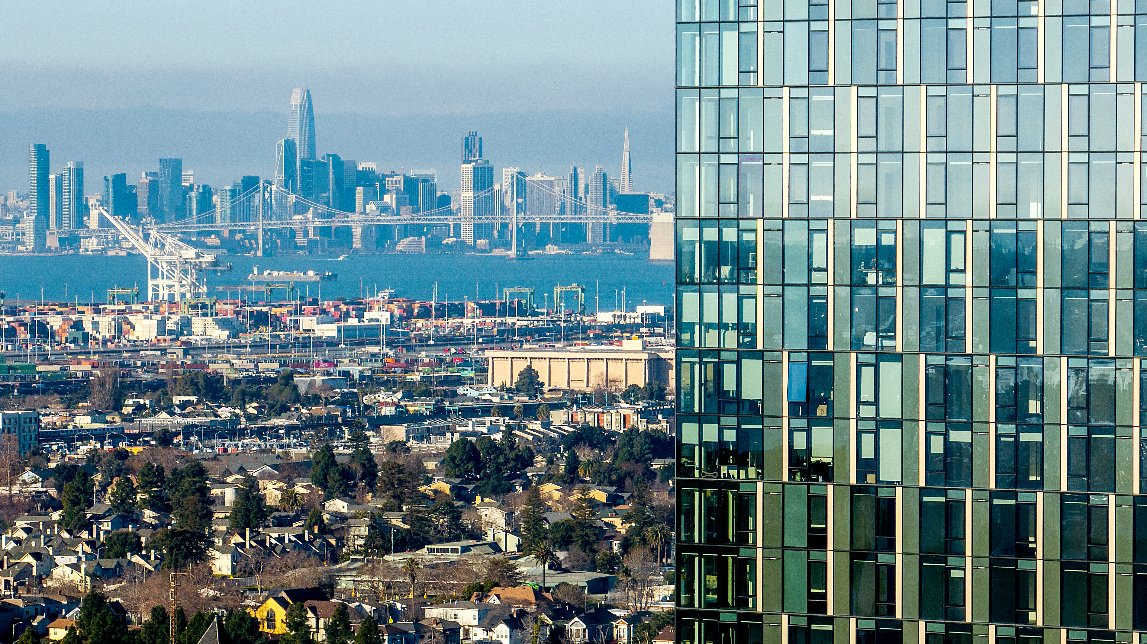 A modern glass building stands in the foreground with a city skyline, a bridge, and shipping containers visible in the background.