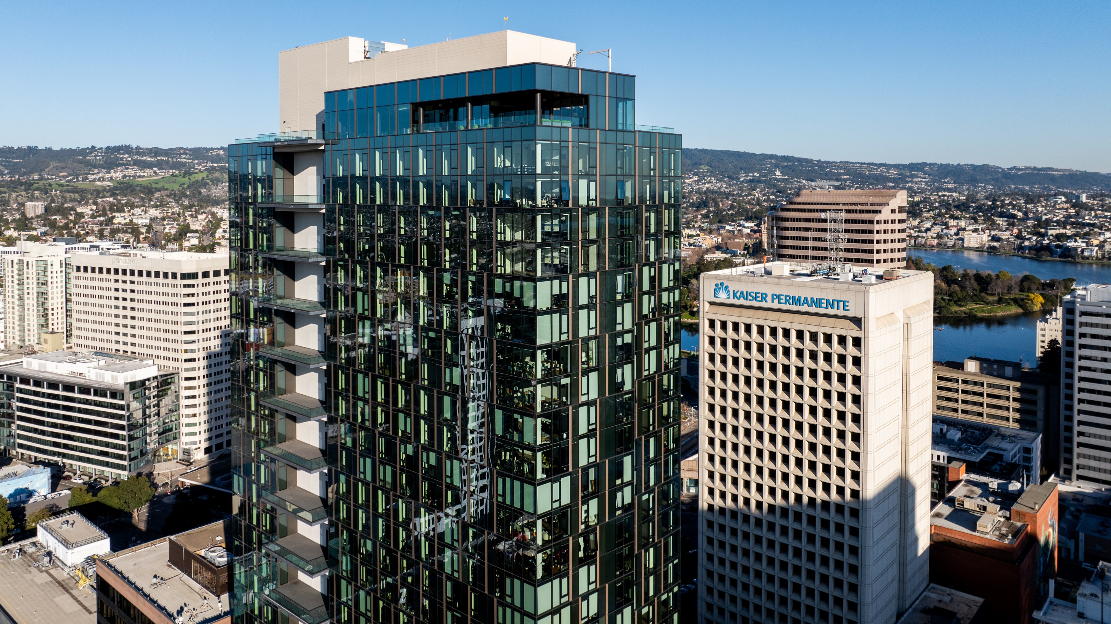 A tall glass skyscraper stands prominently in an urban area with a clear blue sky. Nearby is a white building labeled &quot;Kaiser Permanente,&quot; with a cityscape and water view in the background.